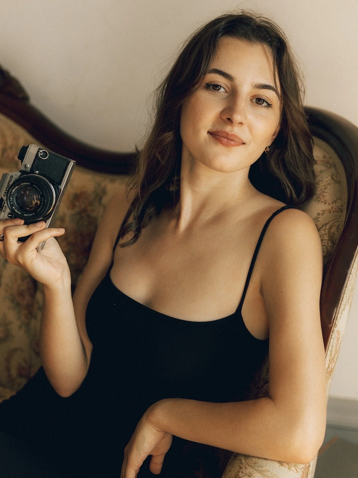 A young woman with long, wavy brown hair and light complexion, wearing a black spaghetti strap top, sitting on a vintage-style sofa with floral upholstery, holding a vintage camera in her right hand, and looking at the camera with a slight smile.