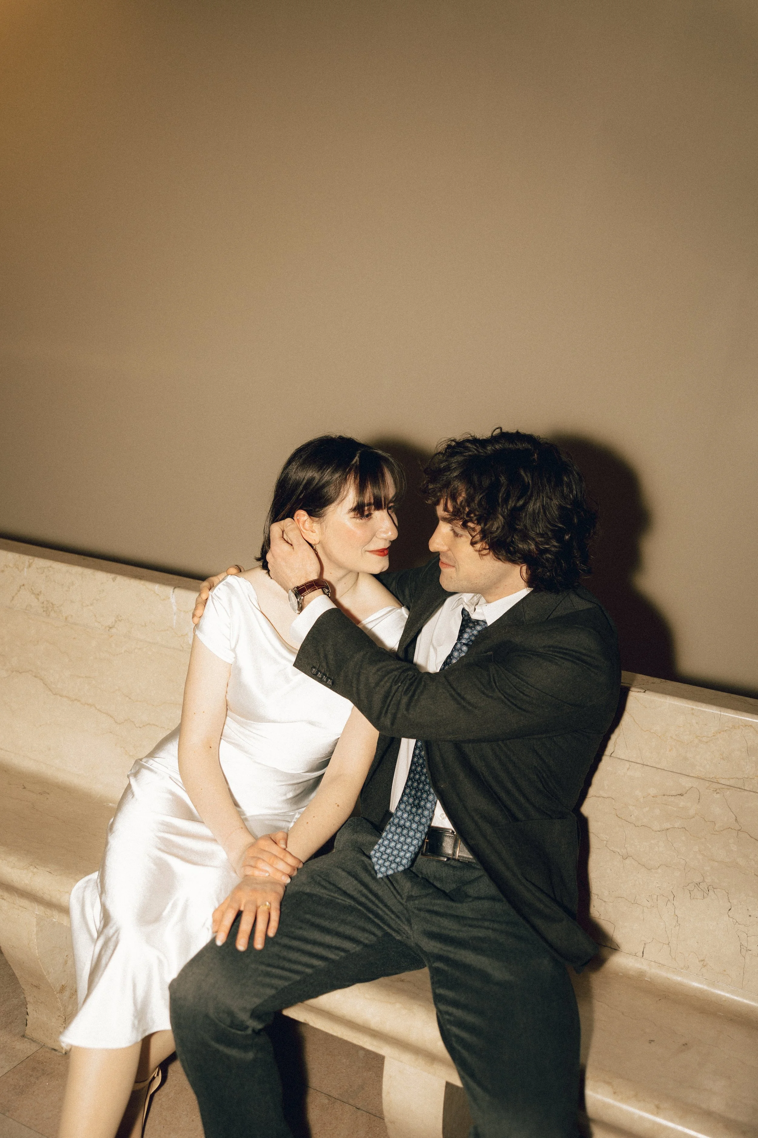 A man and woman sitting closely on a marble bench, gazing into each other's eyes, with the woman's hand gently resting on the man's face.