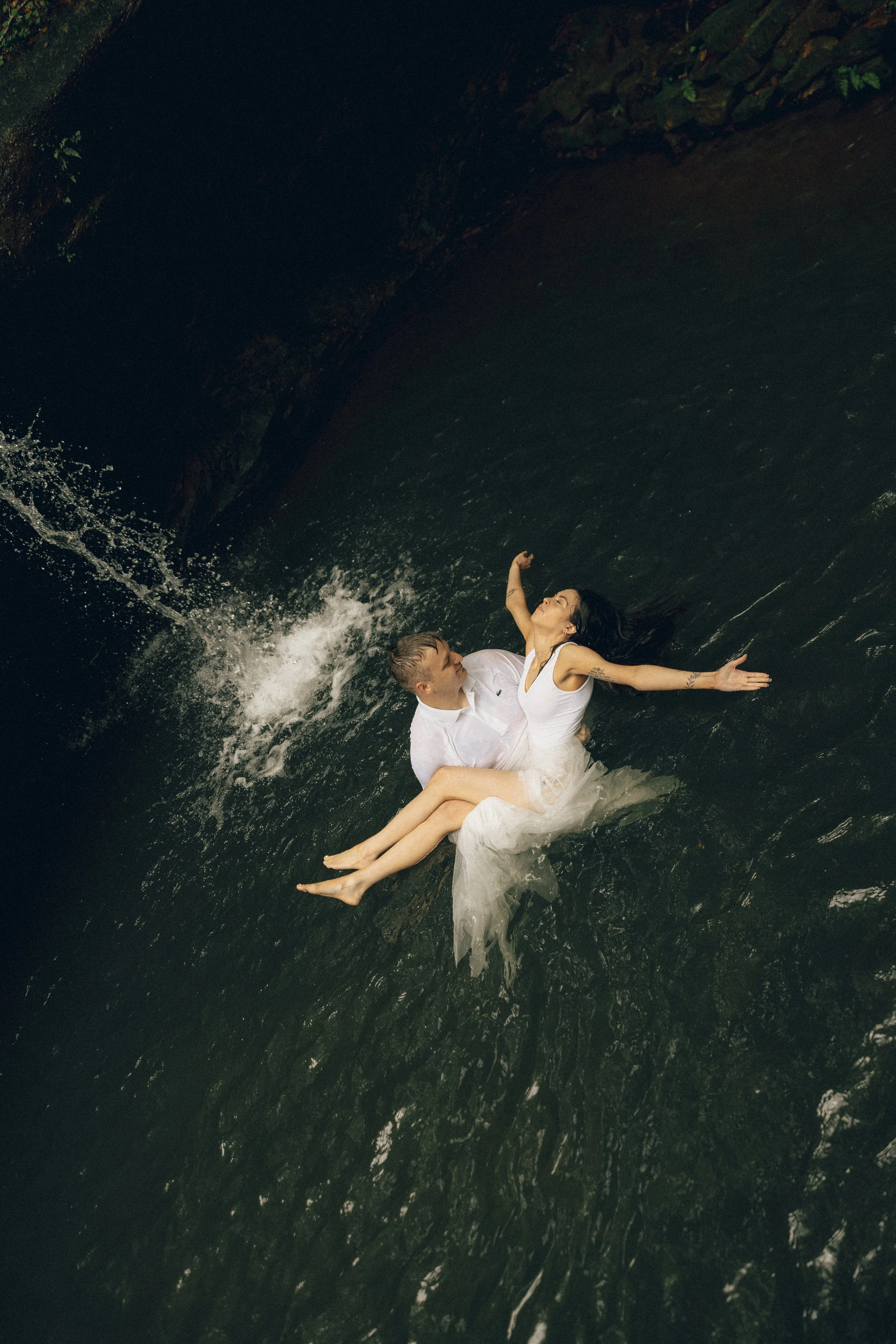 A couple in wedding attire lying on a rock in a river with water splashing around them.