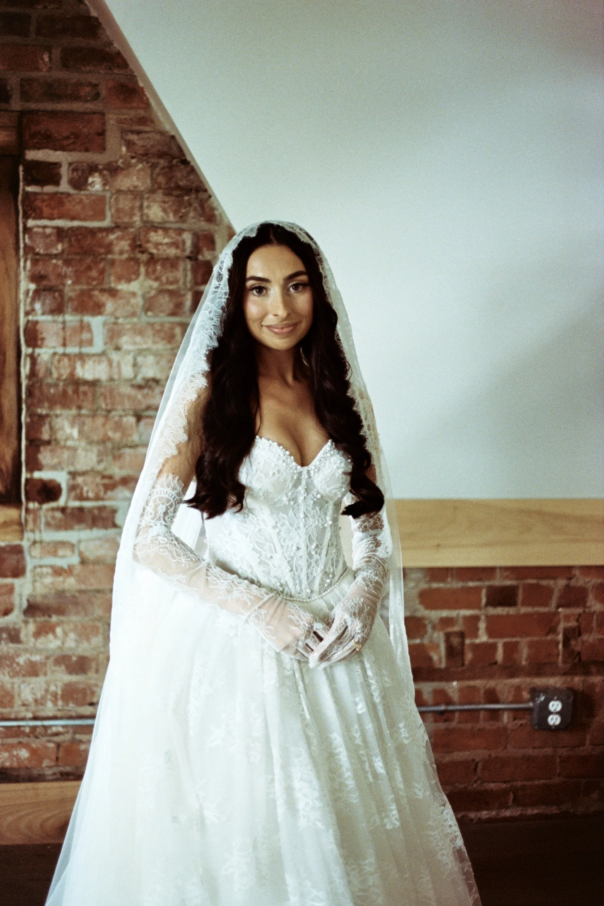 A woman in a white wedding dress with lace details, a veil, and long dark hair smiling inside a room with an exposed brick wall and a wooden surface.