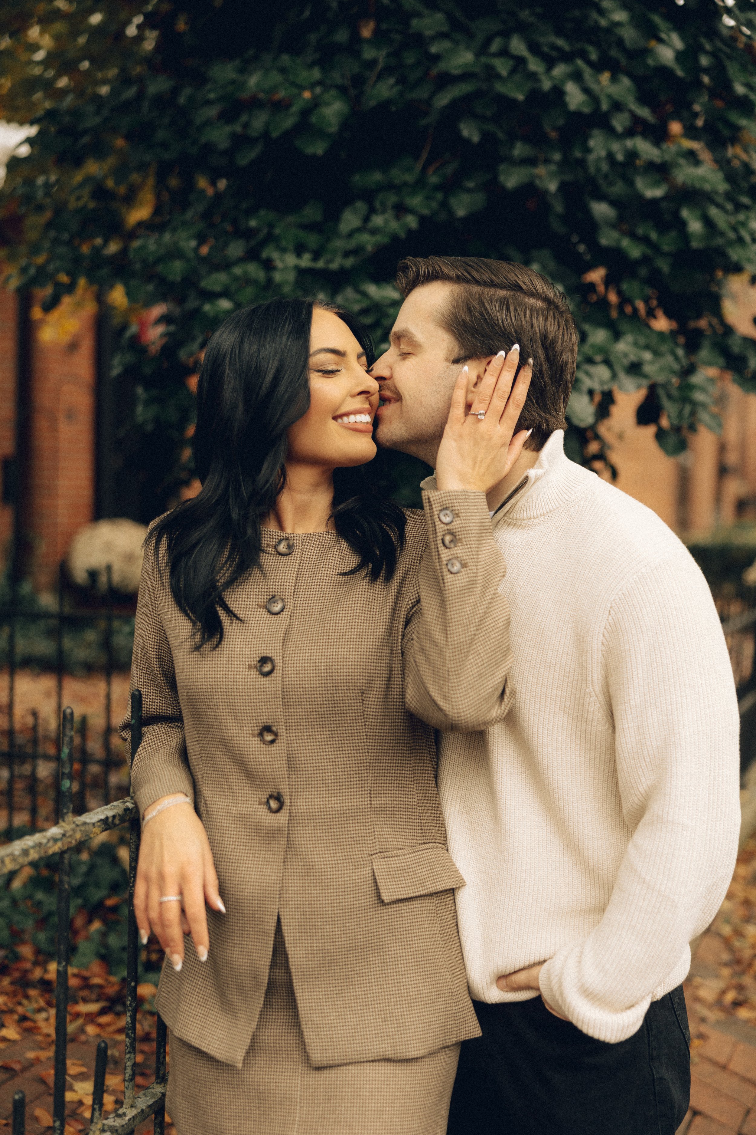 A couple sharing a kiss outdoors, with the woman wearing a checkered coat and the man in a cream sweater, under a tree with green leaves.
