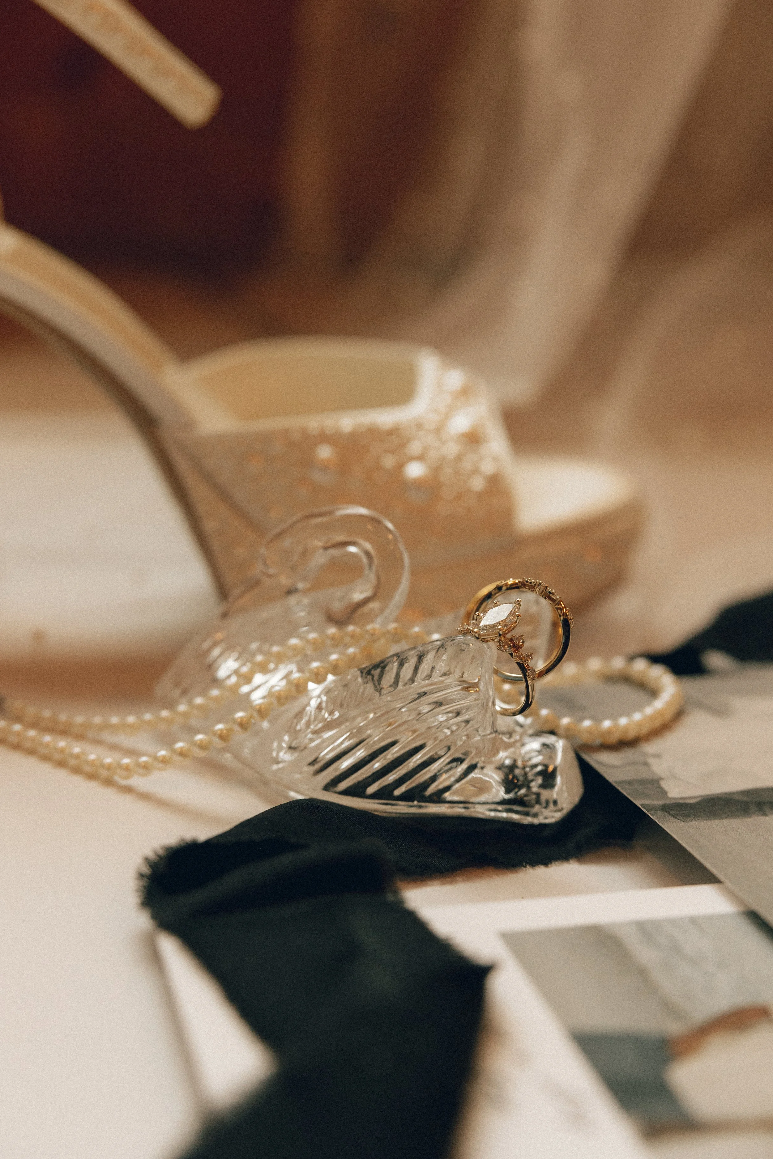 Close-up of jewelry including rings, a pearl necklace, and earrings arranged on glass and fabric, with a blurred background.