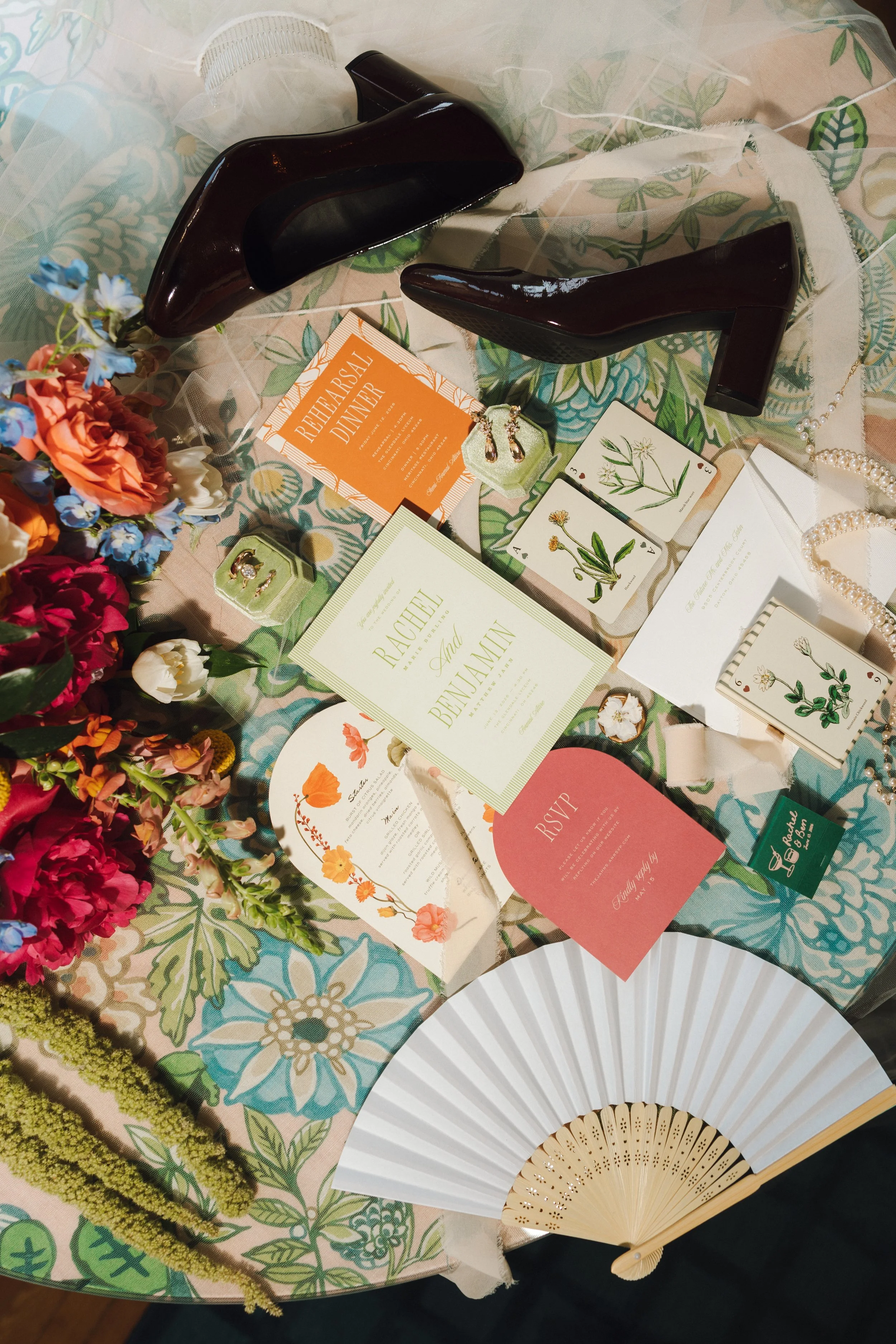A table setup with floral decorations, a hand fan, greeting cards, jewelry, and event invitations for a celebration or wedding.