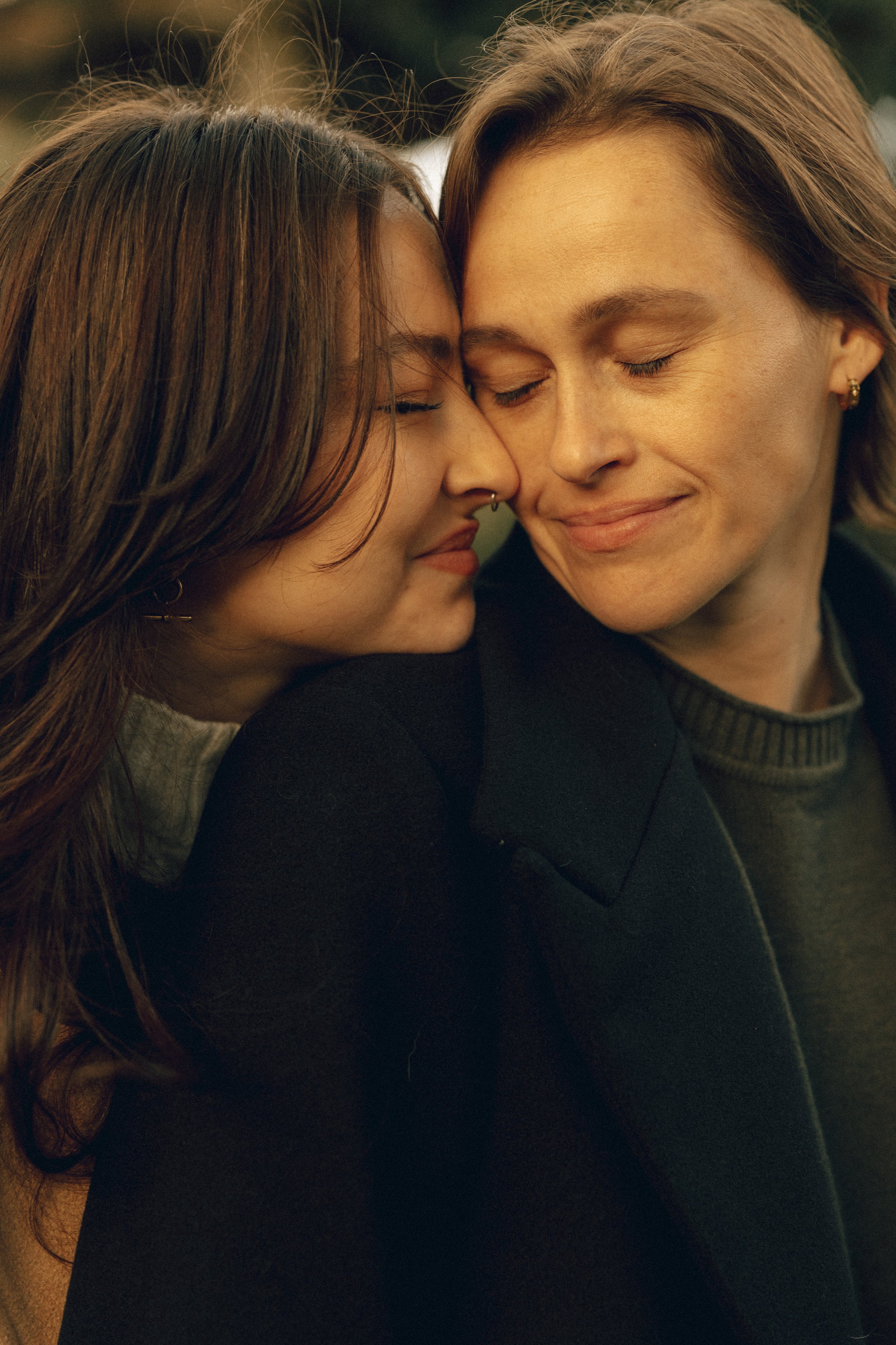 Two women with closed eyes and gentle smiles leaning their foreheads together in an intimate, warm embrace, dressed in dark clothing, outdoors with a blurred background.