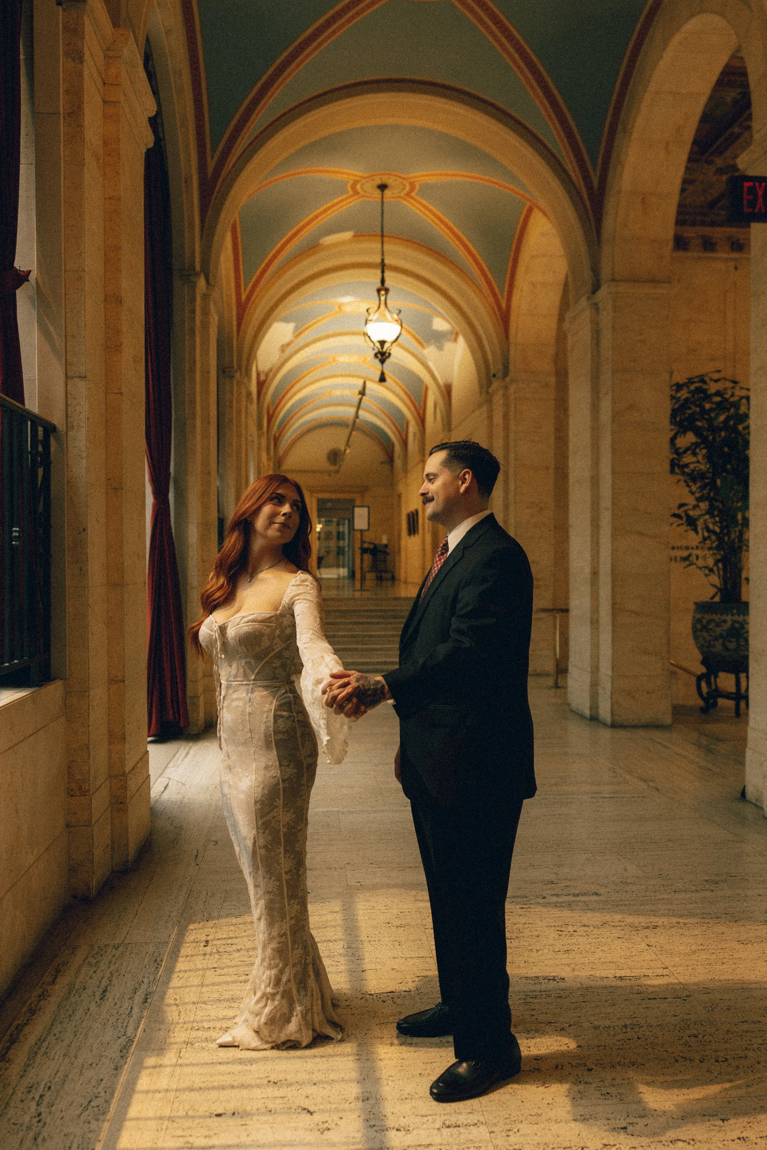 A man and woman holding hands in a grand, arched corridor with ornate ceilings and warm lighting.