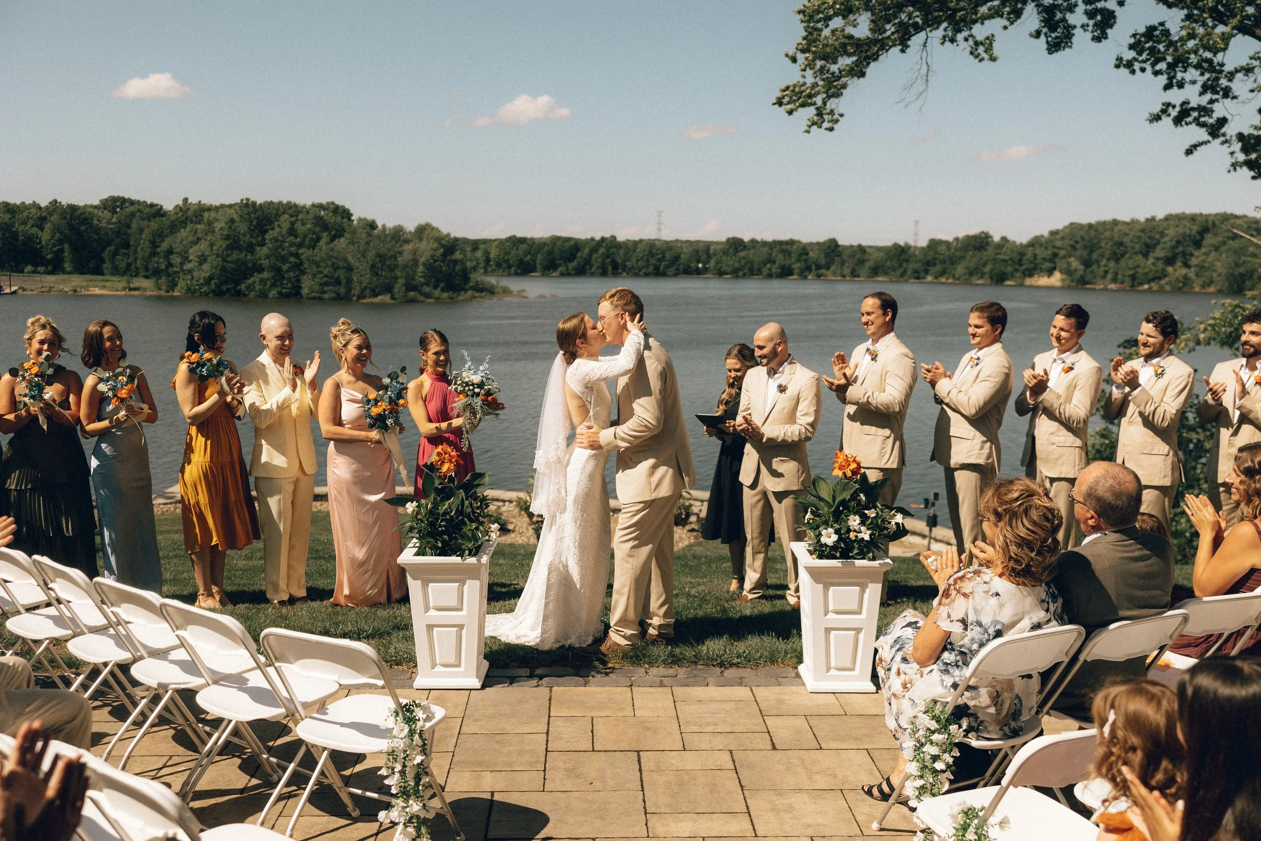A wedding ceremony taking place outdoors by a river with trees in the background. The bride and groom are kissing, surrounded by bridesmaids and groomsmen. Guests are seated in white chairs, clapping and celebrating.