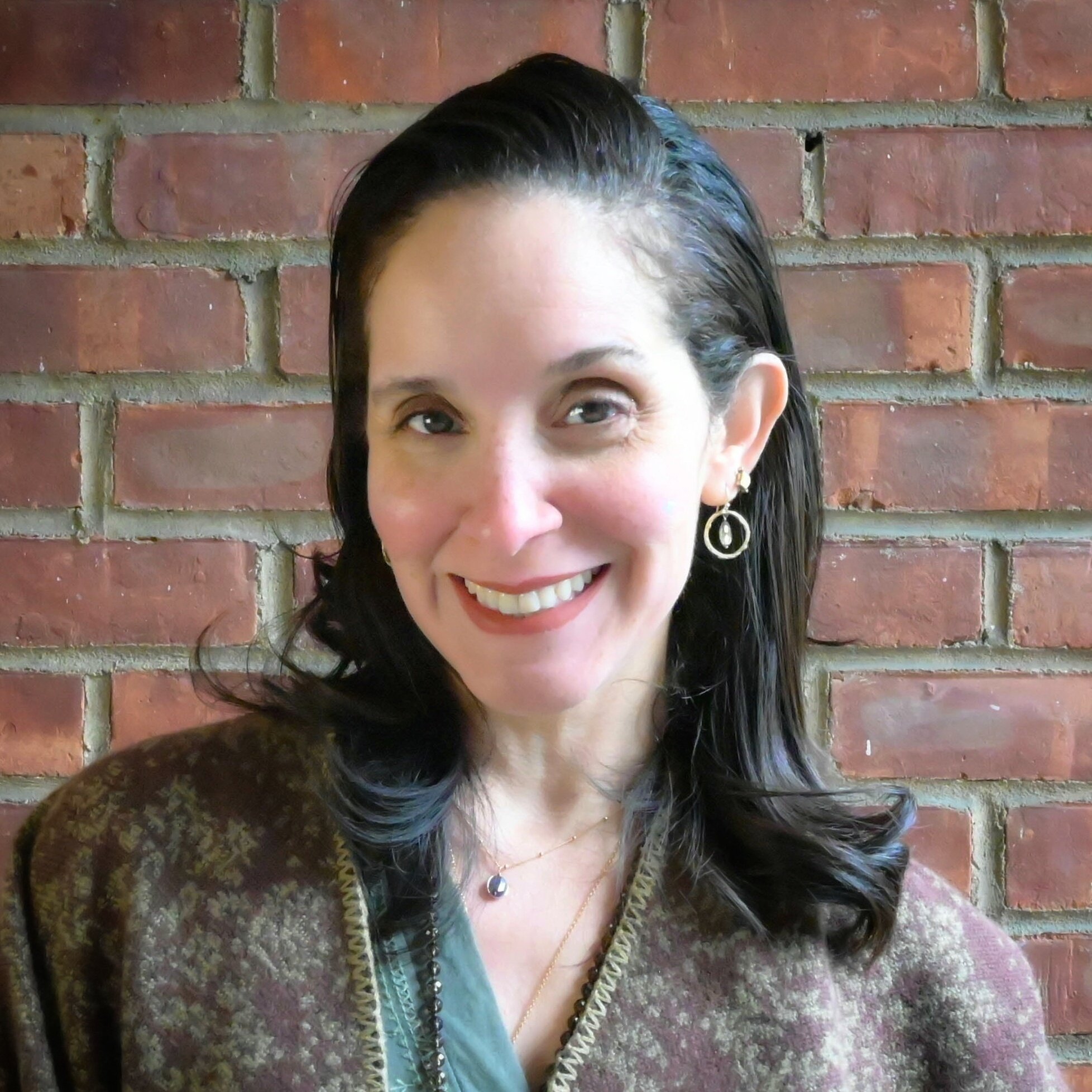 A woman with dark brown hair, smiling, wearing a blue shirt, hoop earrings, and a necklace, with a dark background.