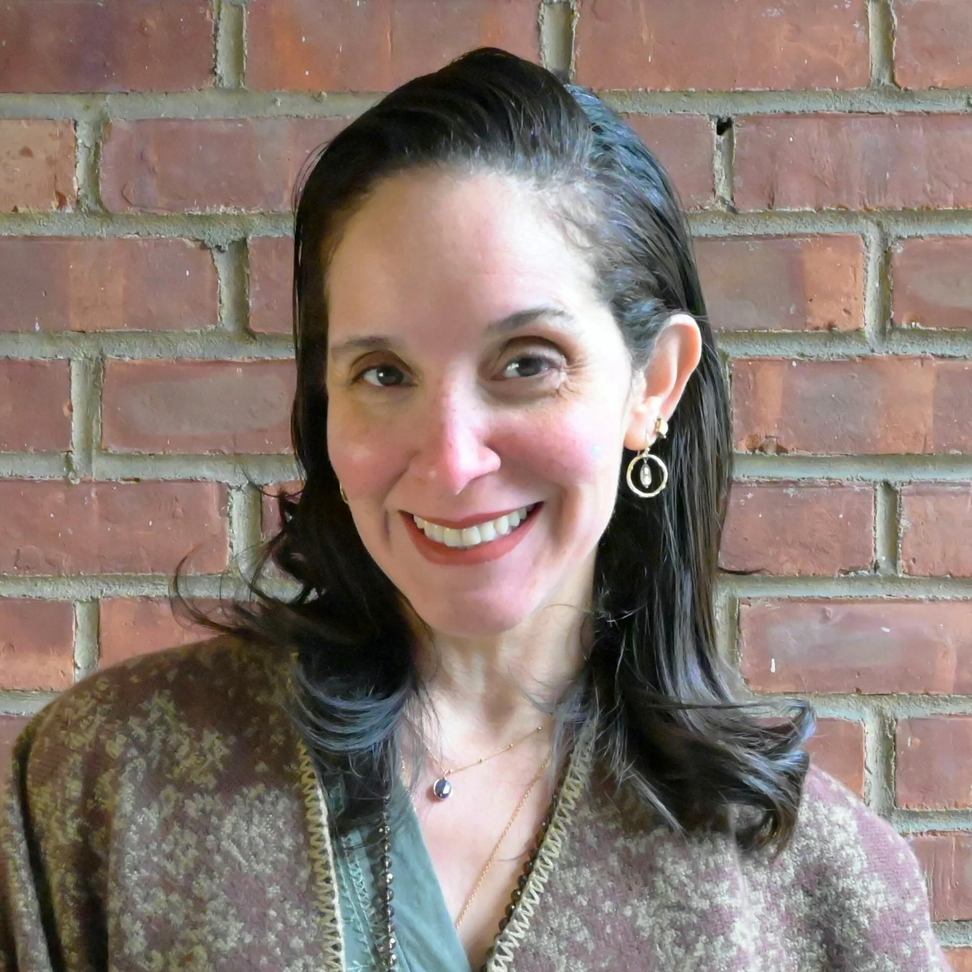 A woman with dark brown hair, smiling, wearing a blue shirt, hoop earrings, and a necklace, with a dark background.