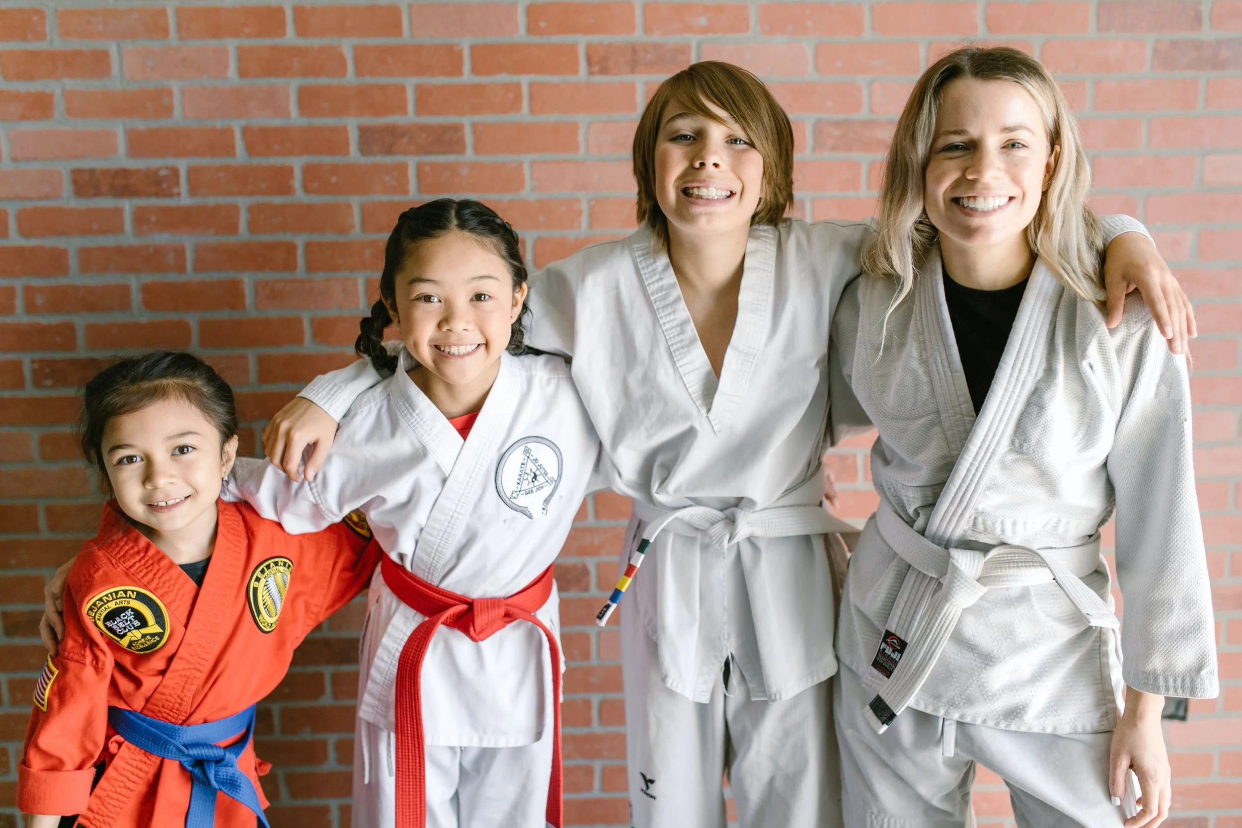 Four young girls in martial arts uniforms smiling and standing arm in arm against a brick wall. They are wearing different colored belts, indicating their martial arts rank.