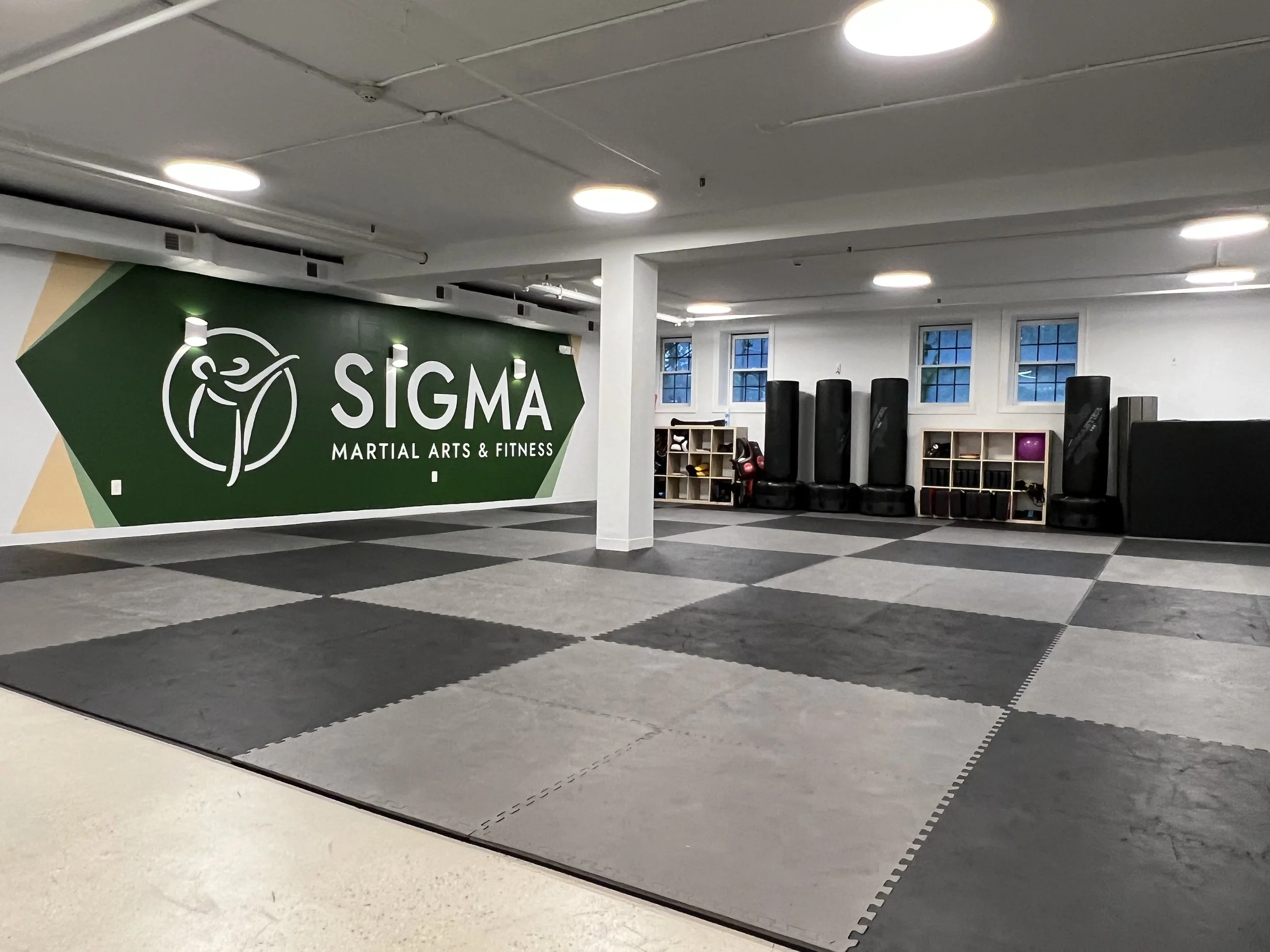 Empty martial arts gym with a large green wall that has the Sigma Martial Arts & Fitness logo, black padded flooring, and equipment such as punching bags, gloves, and storage shelves along the back wall.