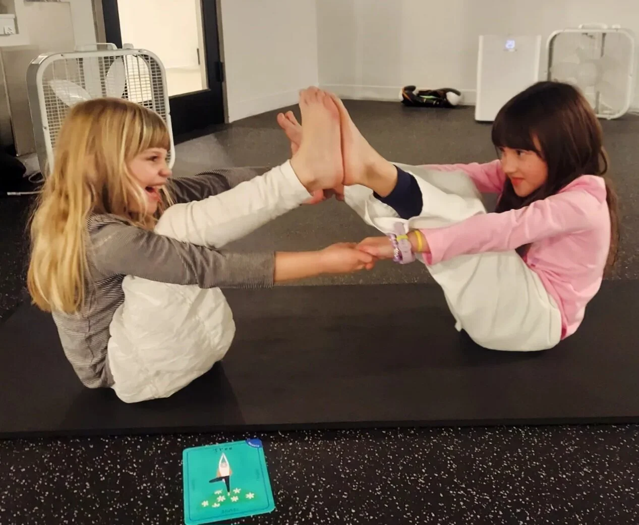 Two young girls are playing a game on a black mat indoors, sitting on their knees and holding hands with their legs extended upward, touching soles together. They are smiling and facing each other, engaging in playful activity.