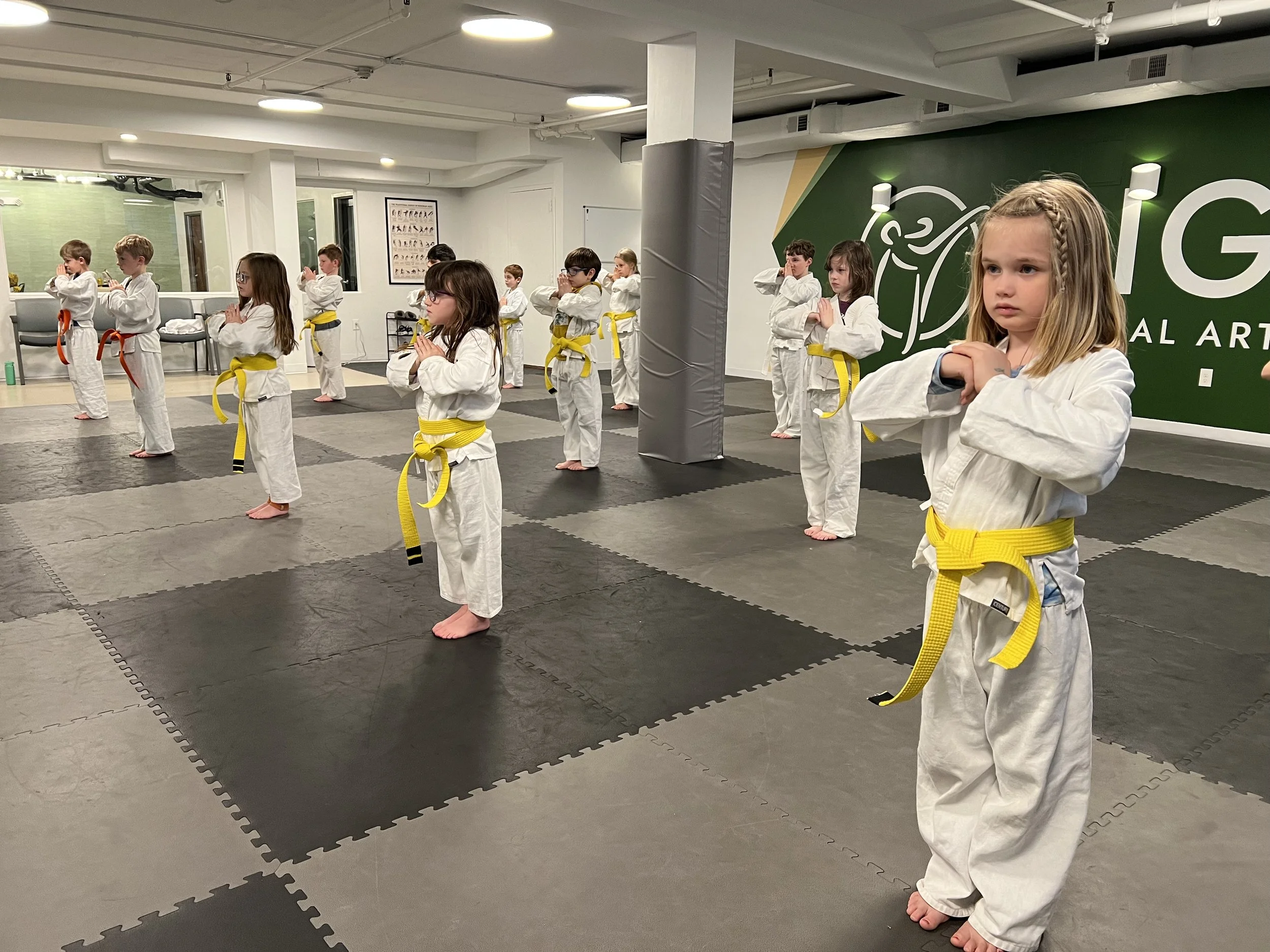 Children in white martial arts uniforms practicing martial arts in a dojo, standing in formation with yellow belts, in a training room with padded floors and a green wall with a logo.