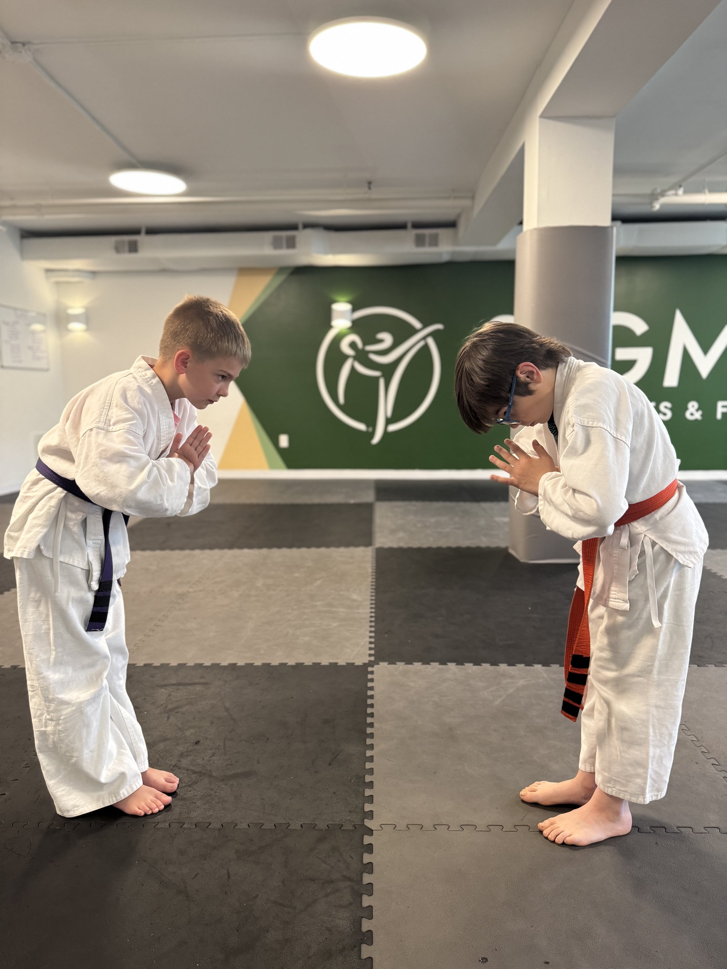 Two young boys in martial arts uniforms bowing to each other during a class in a gym.