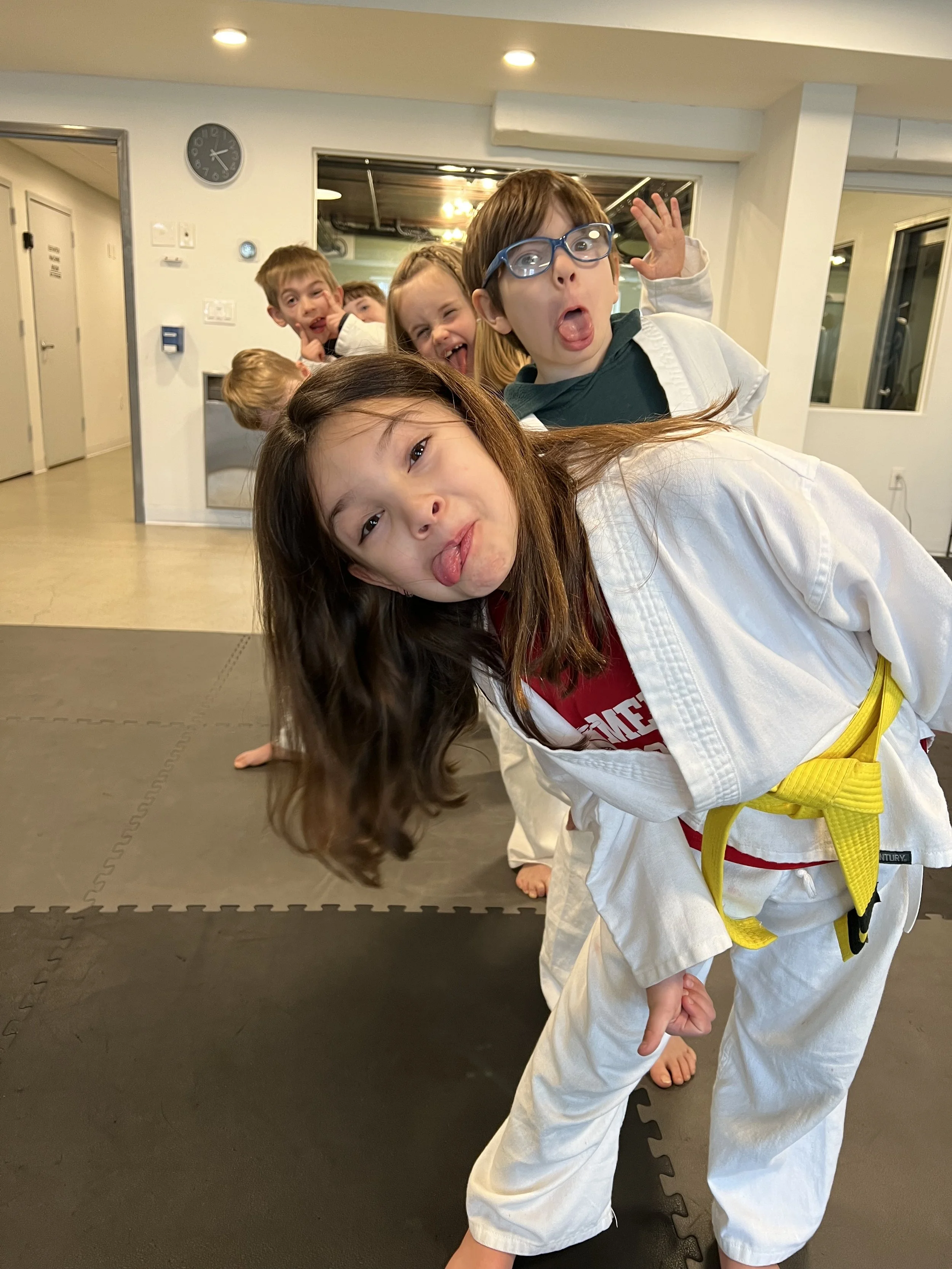 Children in martial arts uniforms making playful faces and sticking out their tongues, practicing a pose in a dojo or martial arts studio.