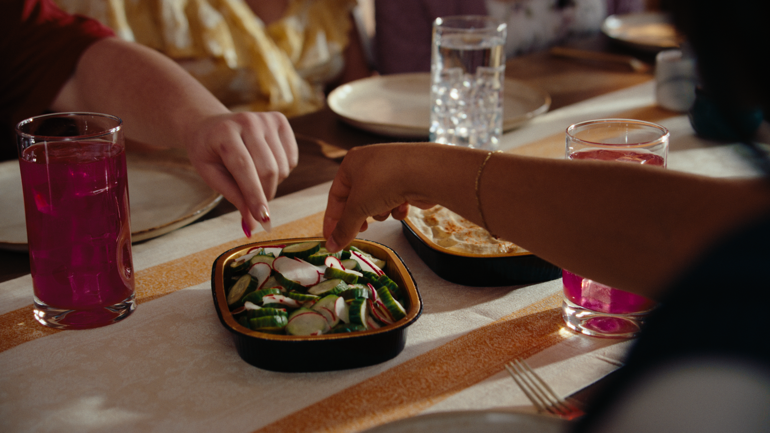 Close up of a dining table with two hands reaching into a dish of Brassica cucumbers and radishes. A dish of hummus and glasses filled with Brassica lemonade sit nearby. The still is from "wow," a narrative food commercial produced by waxpoet, a comm