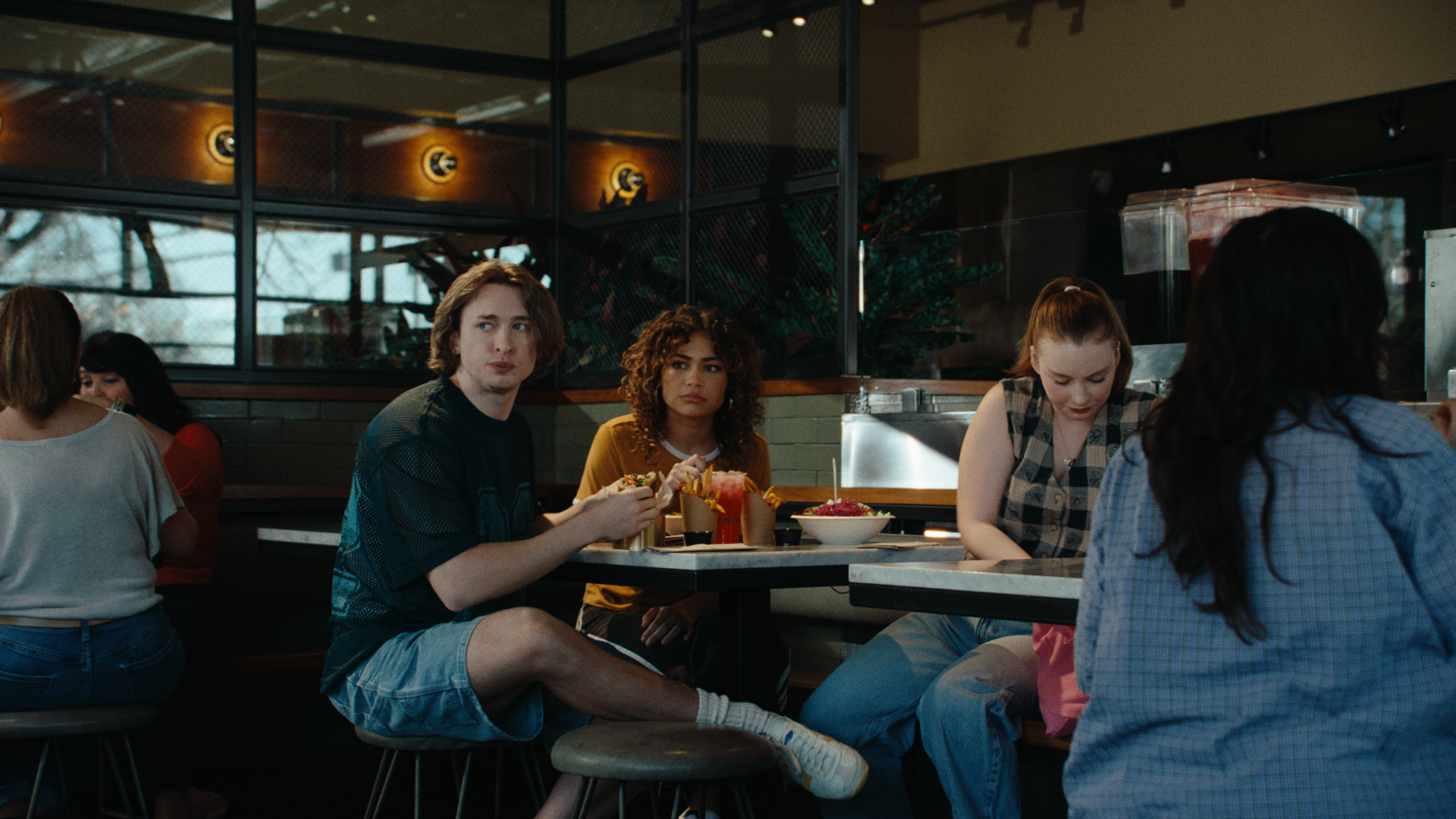 A group of friends sit at a table in a Brassica restaurant, surrounded by other patrons. Two of the friends are turned toward the camera and looking out of a window, while the other is looking through a bag. The still is from "let's stay," a food com