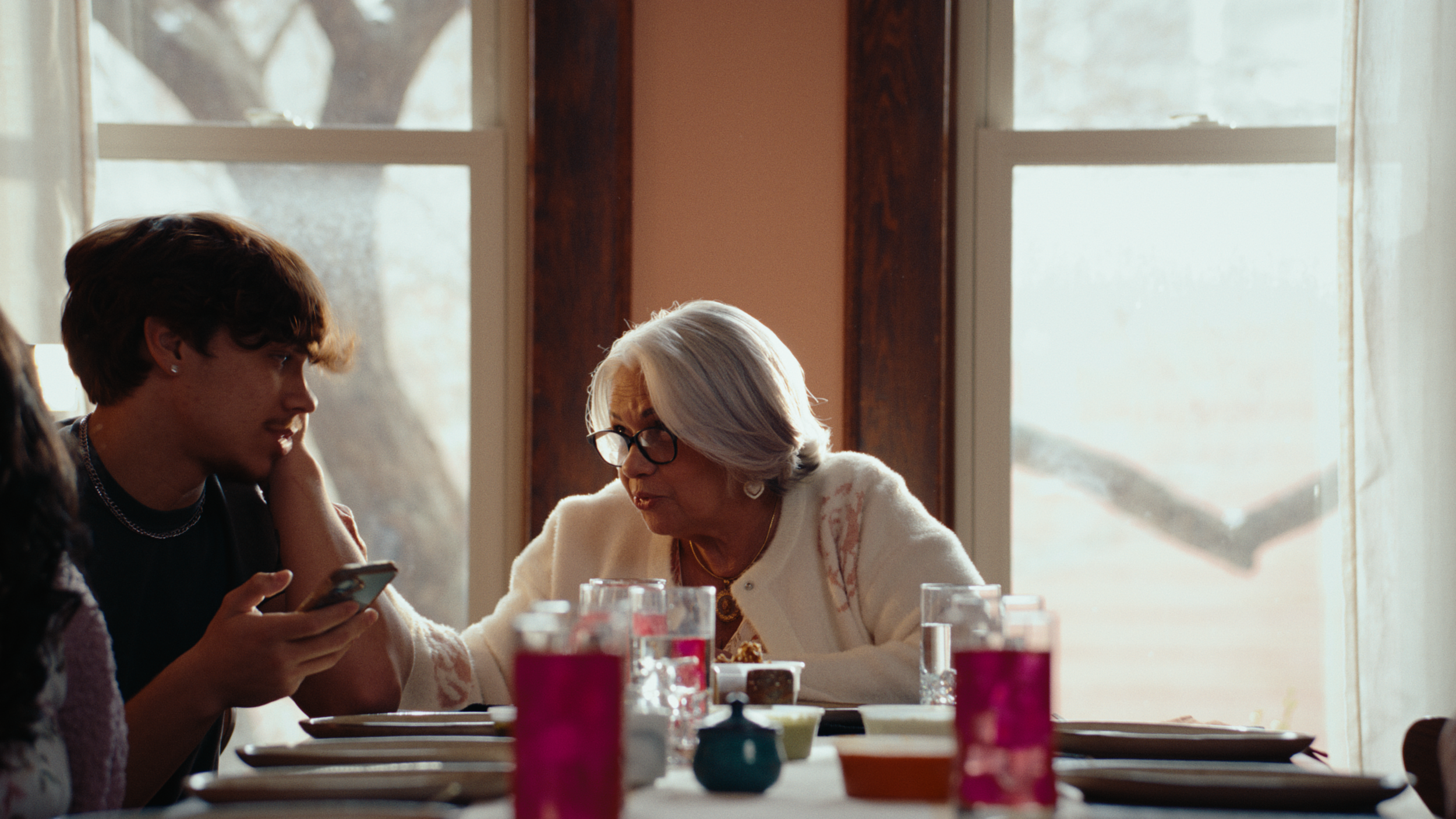 An elderly woman and a young man converse at a dining table set with empty plates and glasses filled with Brassica's pink lemonade, with windows in the background. The still is from "wow," a narrative food commercial produced by waxpoet, a commercial