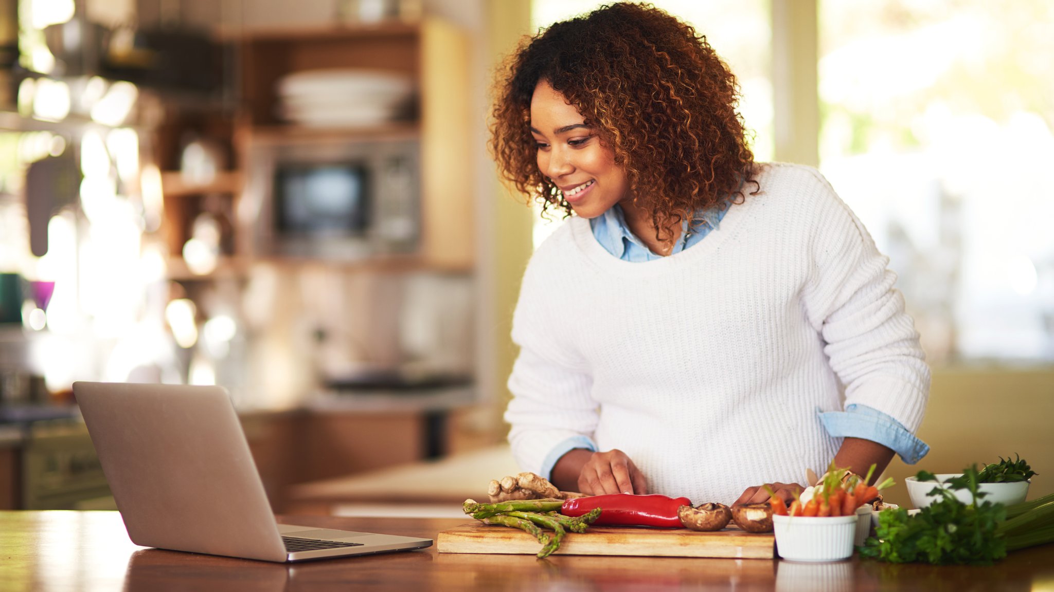 A woman smiling at her laptop with ingredients laid out on the table in front of her