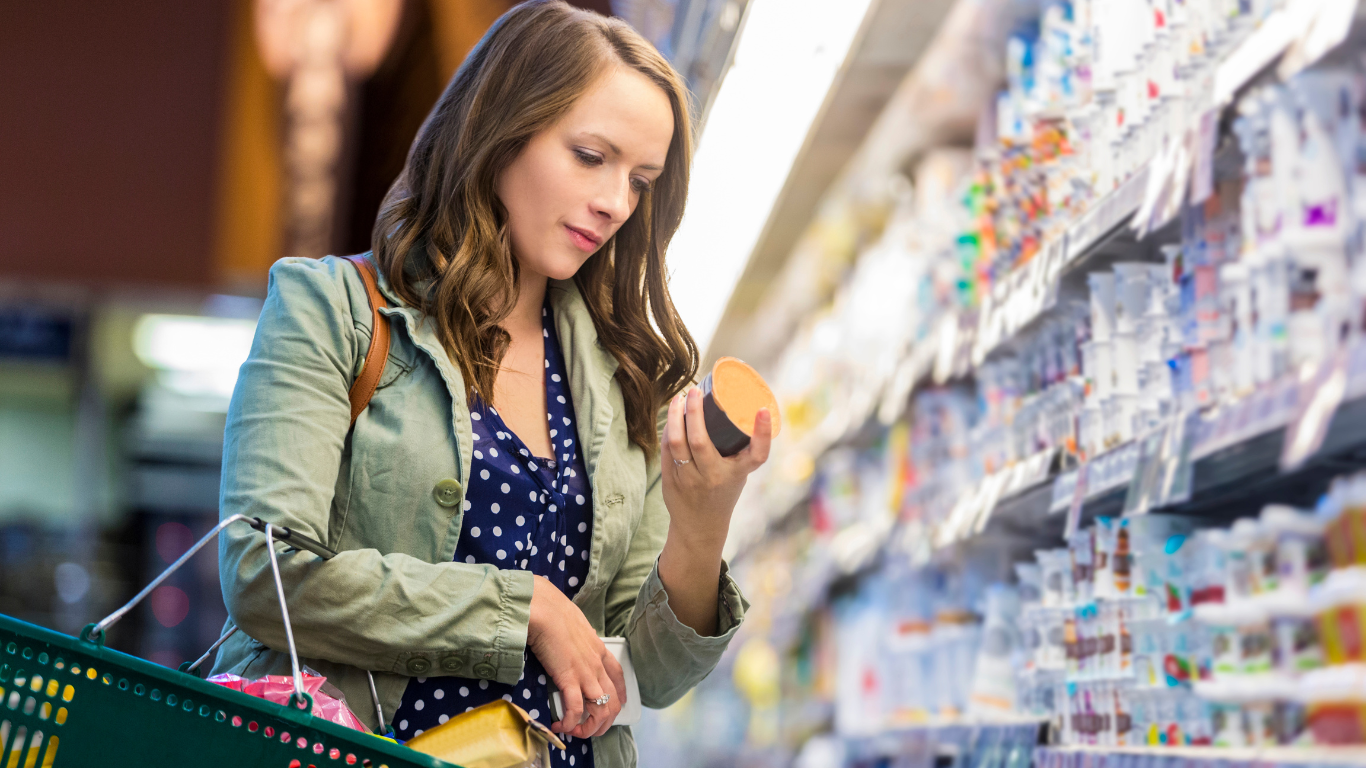 A woman in the grocery store carrying a basket and holding a yogurt while reading the nutritional facts