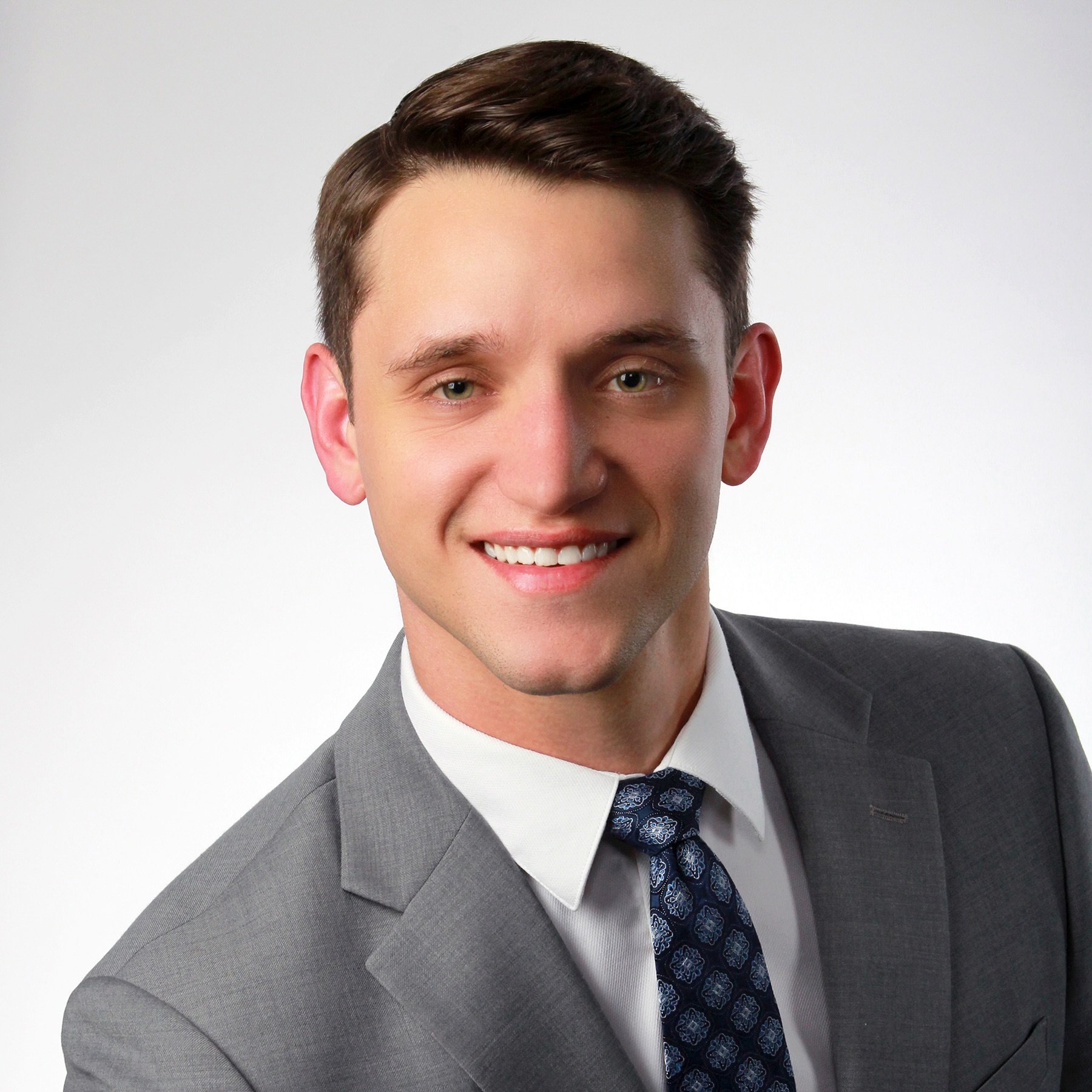 Portrait of a young man in a gray suit with a white shirt and patterned blue tie, smiling with white teeth, against a plain white background.
