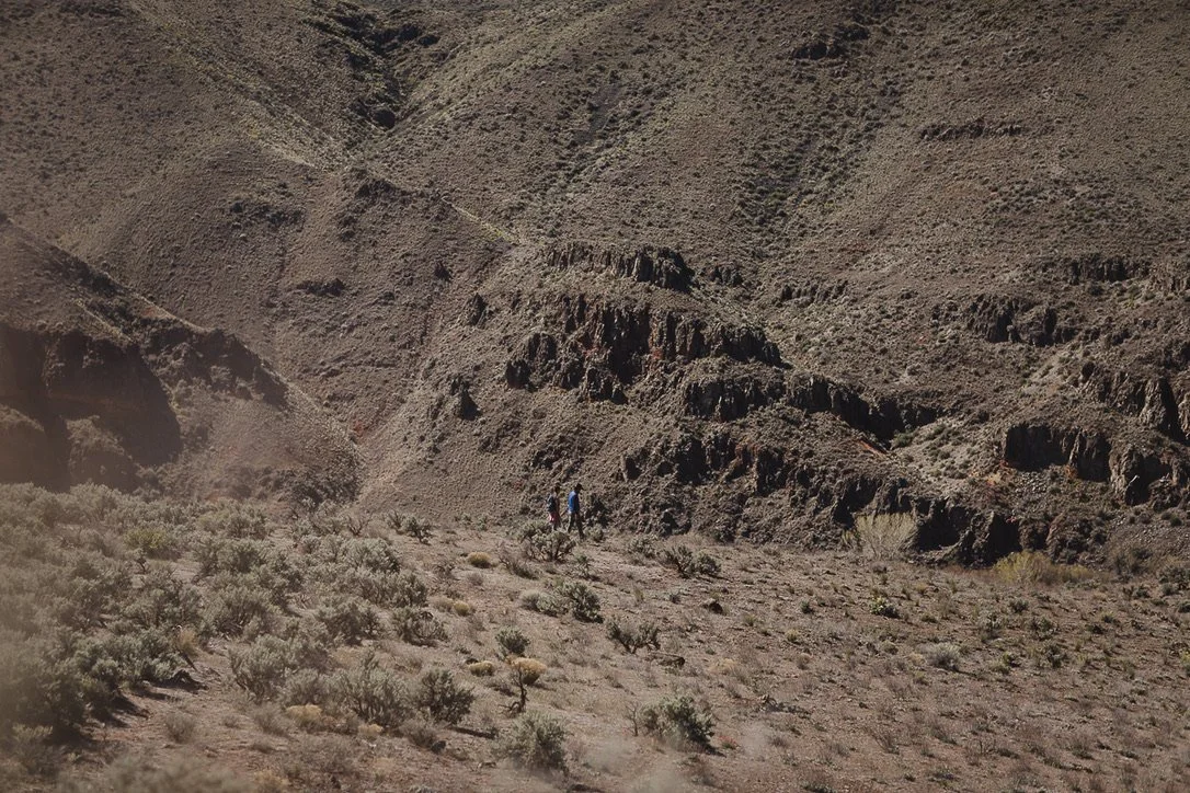 Three people hiking in a dry, mountainous desert landscape with sparse vegetation and rocky terrain.