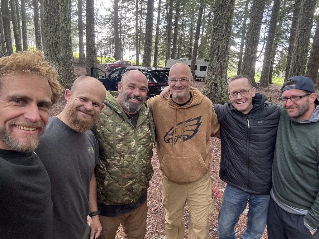 Six men standing in a forest, smiling with their arms around each other's shoulders, with parked cars and trees in the background.