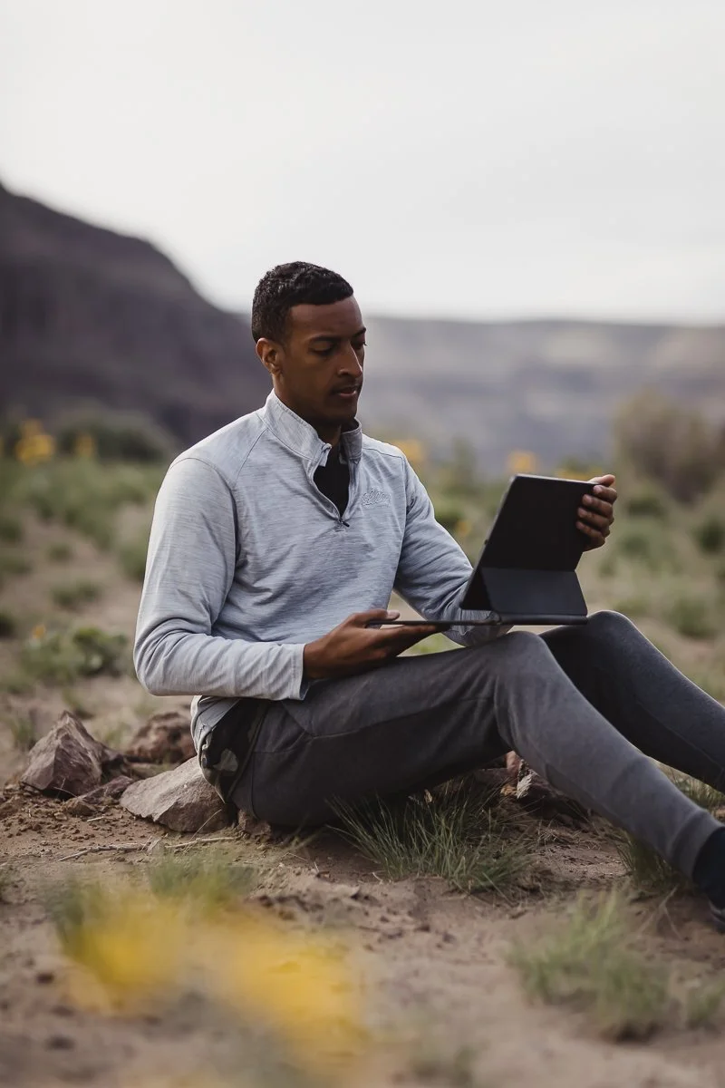 A young man sitting outdoors on rocky terrain, holding a tablet and looking at it, with a scenic landscape of hills and plants in the background.