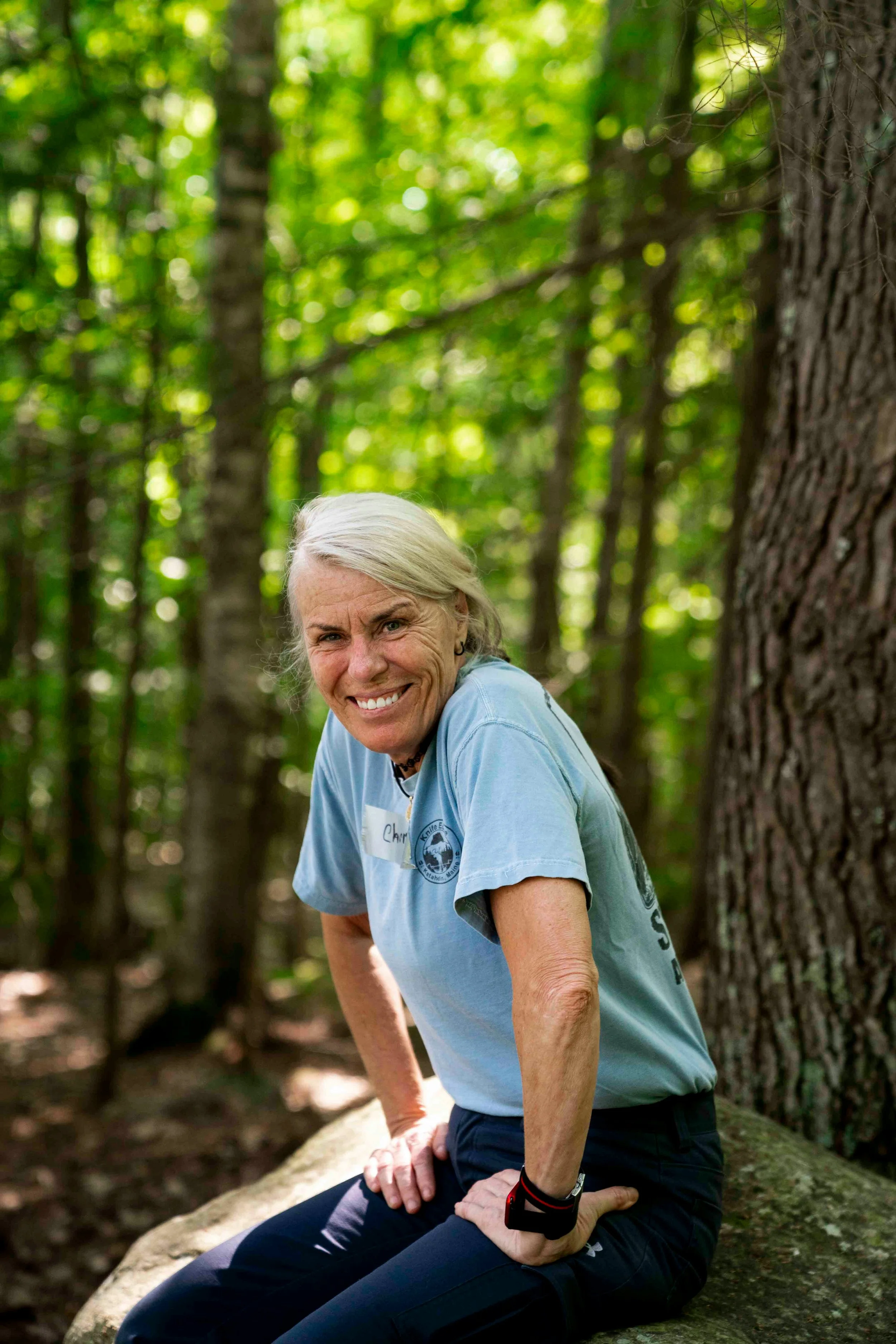 An elderly woman with gray hair smiling while sitting on a large rock in a wooded area, wearing a light blue t-shirt and athletic pants.