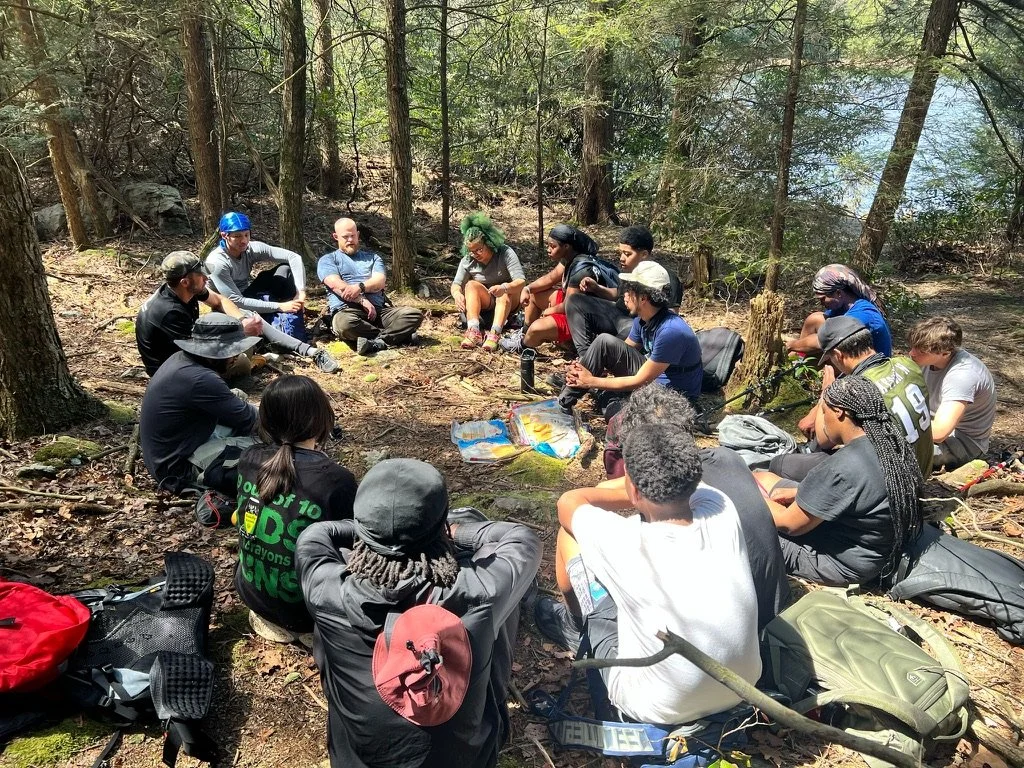 Group of people sitting in a circle in a wooded forest area, some with backpacks and outdoor gear, during daytime.