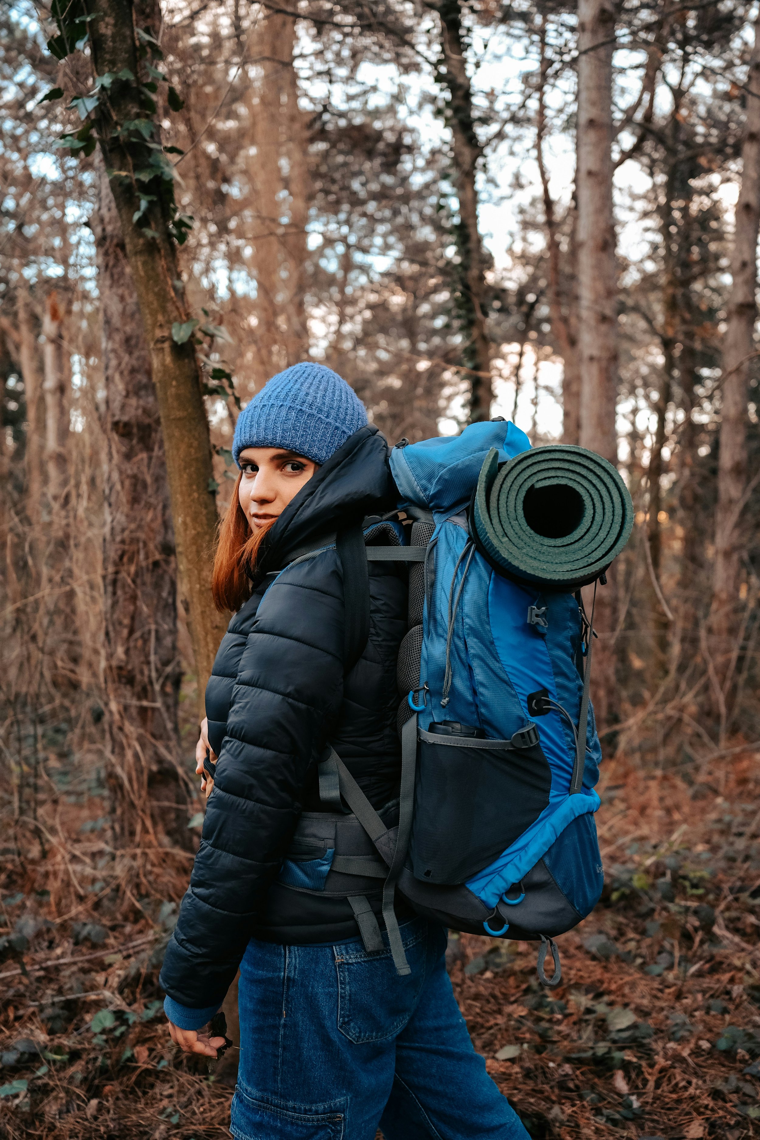 A woman with long hair wearing a blue knit hat and a black puffy jacket, standing in a forested area during fall or early winter. She is carrying a large blue backpack with a rolled-up yoga mat attached on top.