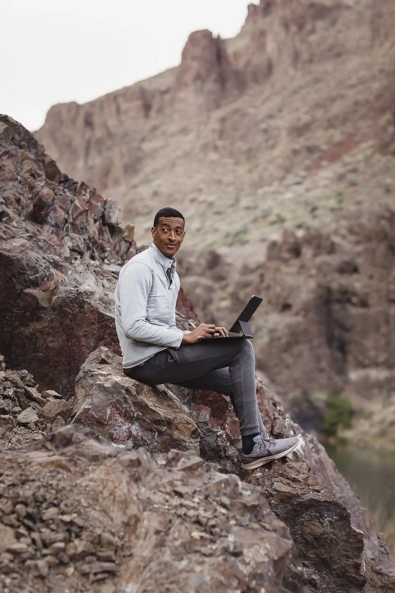 A man sitting on a rocky hillside working on a laptop with a canyon in the background.