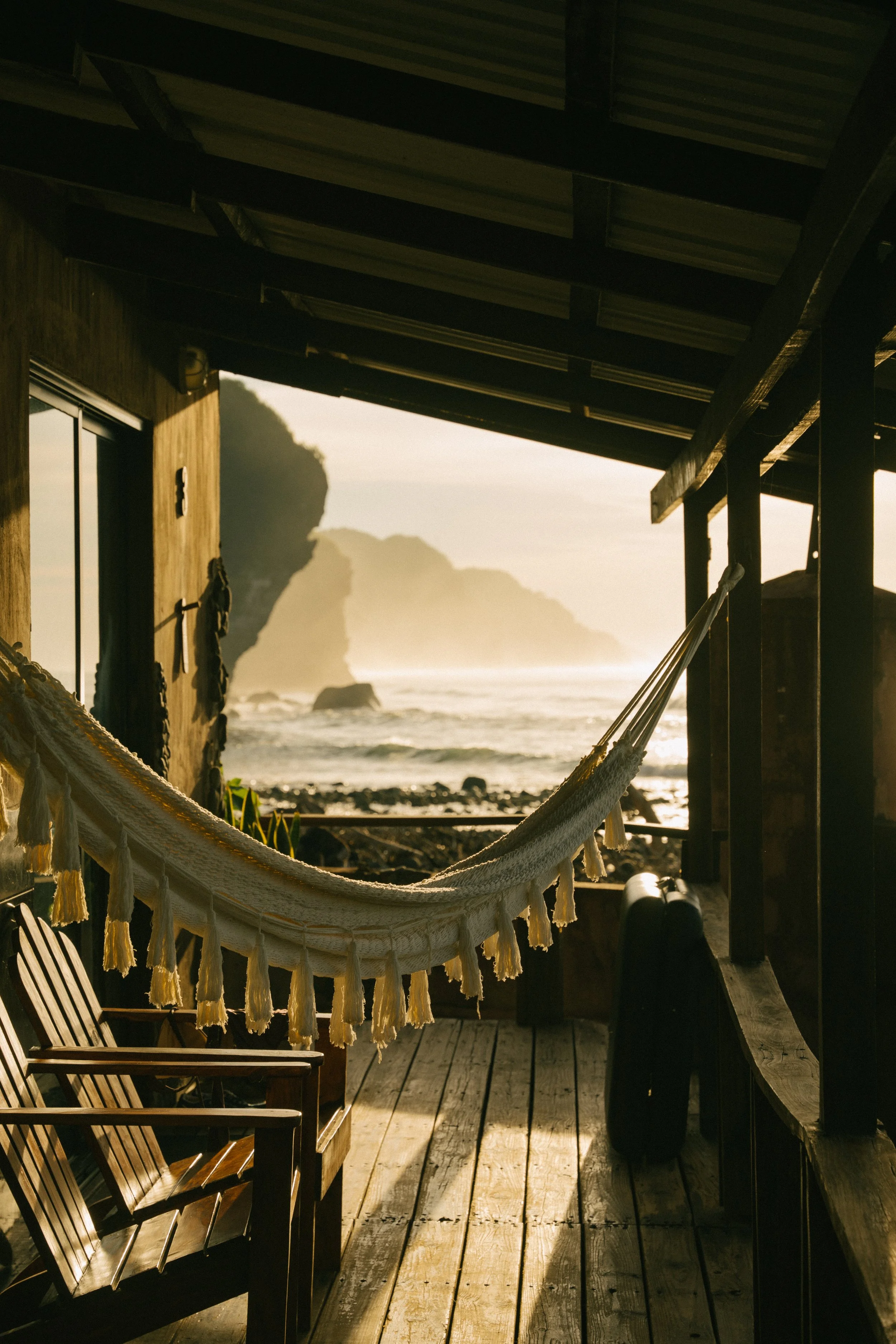 A porch with wooden chairs, a hammock, and a view of the beach and rocky cliffs during sunset.