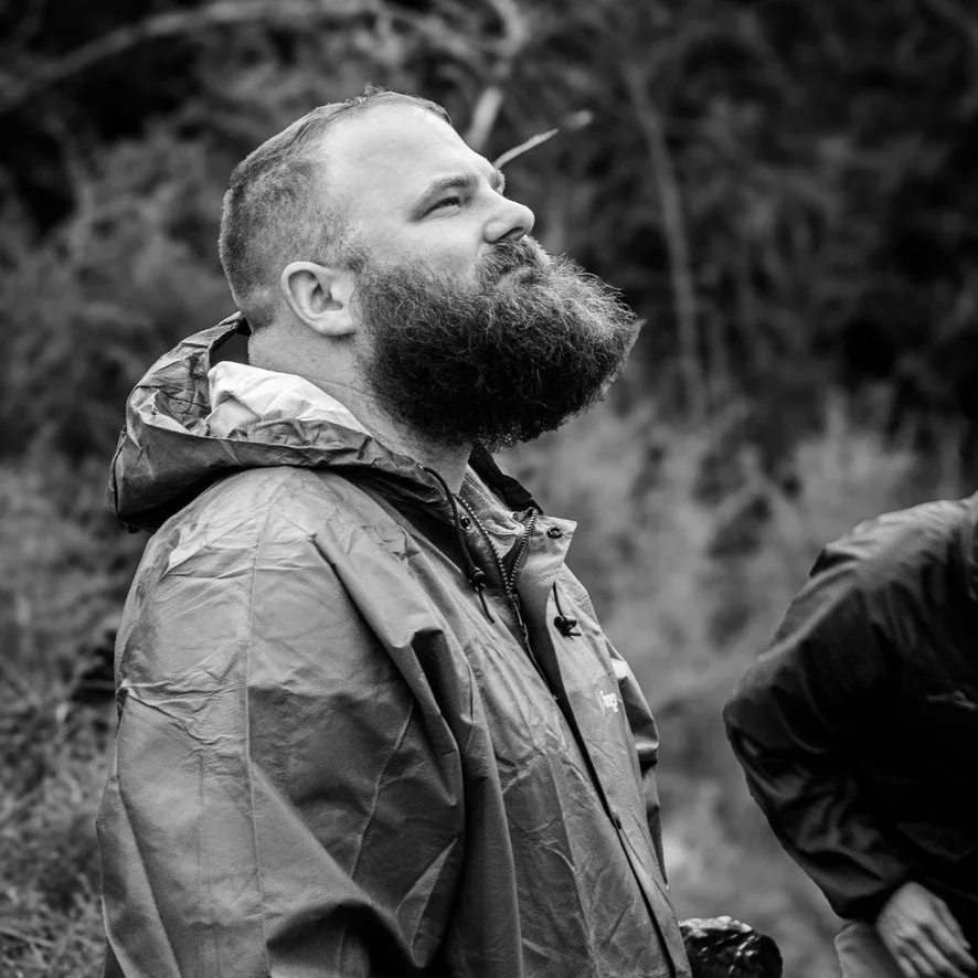 A man with a beard wearing a rain jacket, looking upward, outdoors in a forested area.