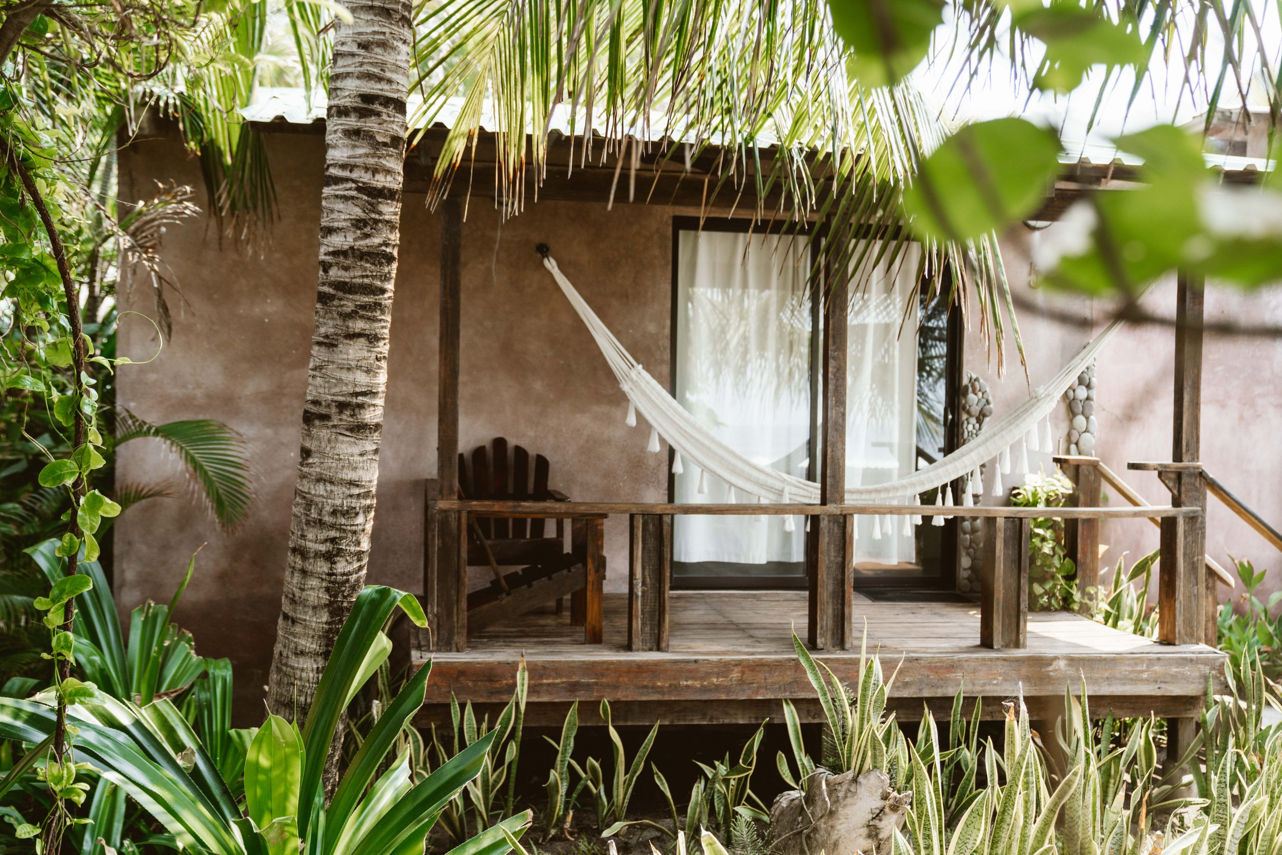 A wooden balcony with a white hammock, a chair, and plants surrounding a small house or cabin. Tall palm and leafy trees shade the area.