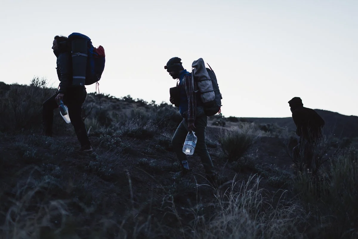 Three hikers with backpacks and water bottles hiking up a hill at dusk or dawn.