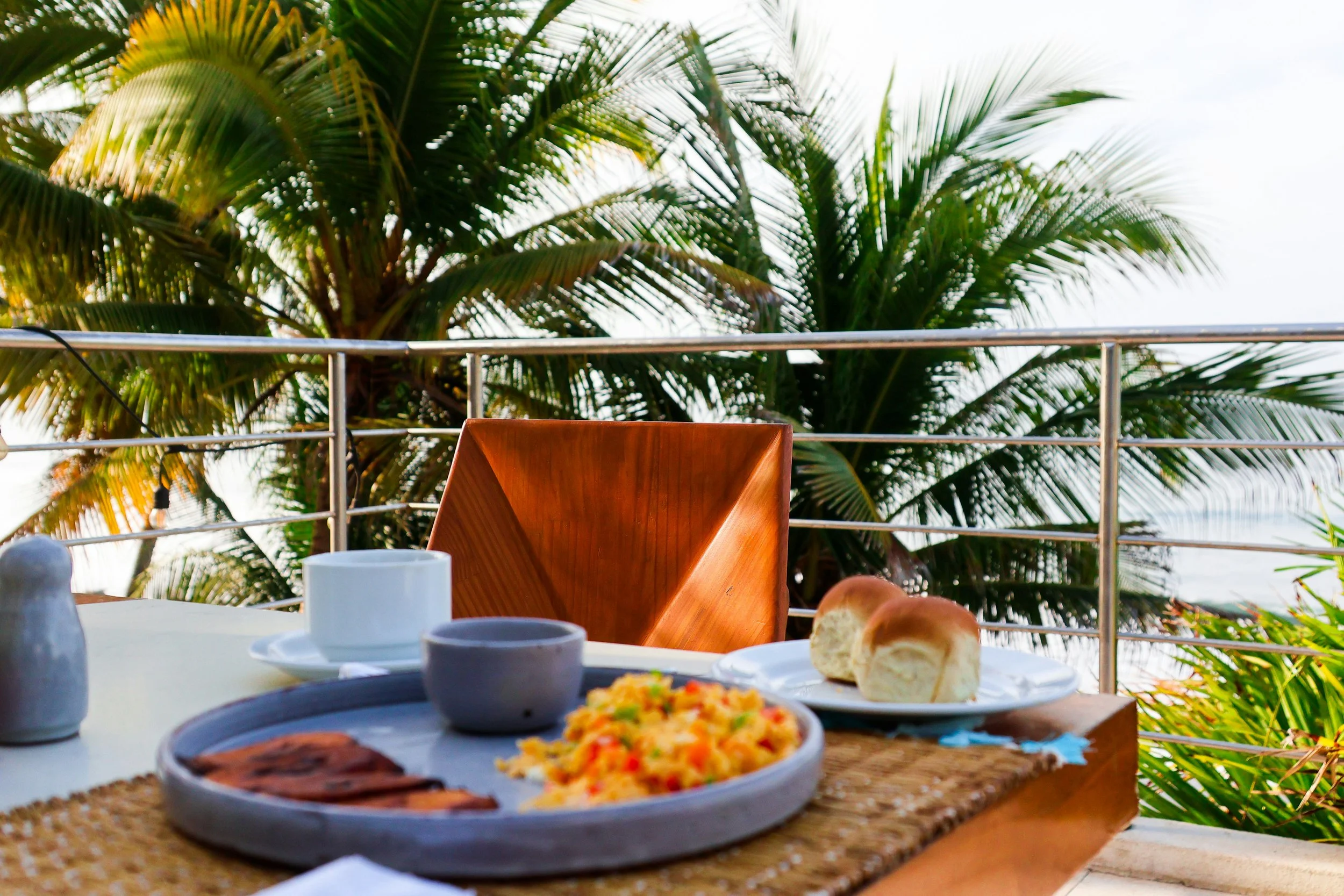 A table with plates of food including rice, bread rolls, and a meat dish, set outdoors on a balcony with tropical palm trees in the background.