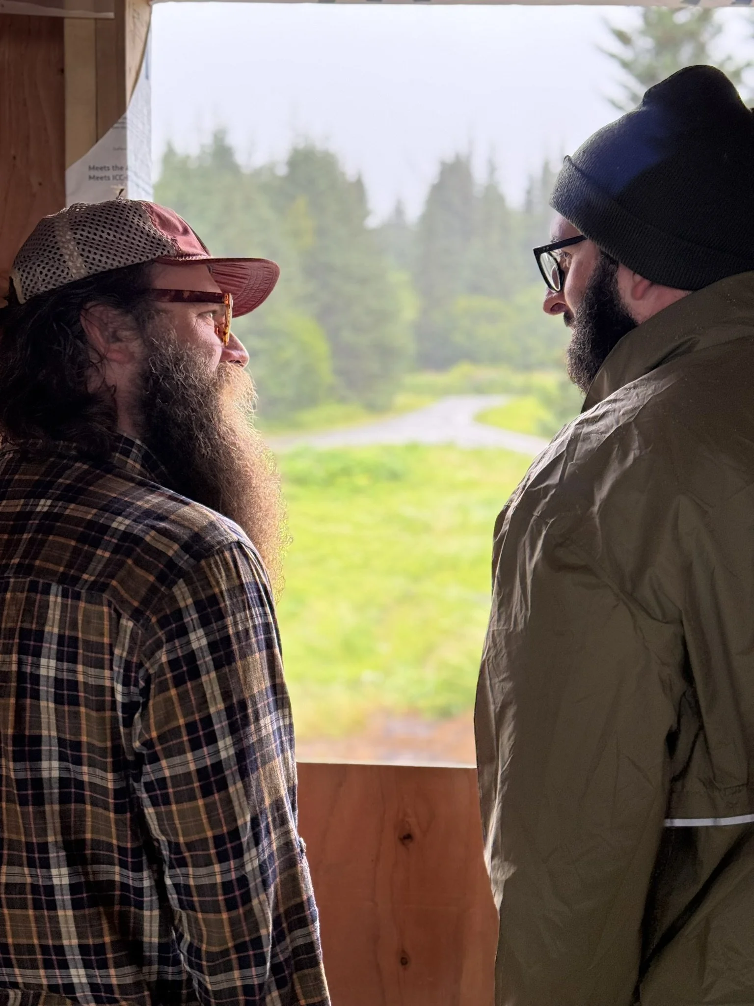 Two men standing face-to-face by a window with a view of green trees outside, engaging in conversation. The man on the left wears a plaid shirt, a cap, and glasses, with a long beard. The man on the right wears a cap, glasses, and a jacket, also with