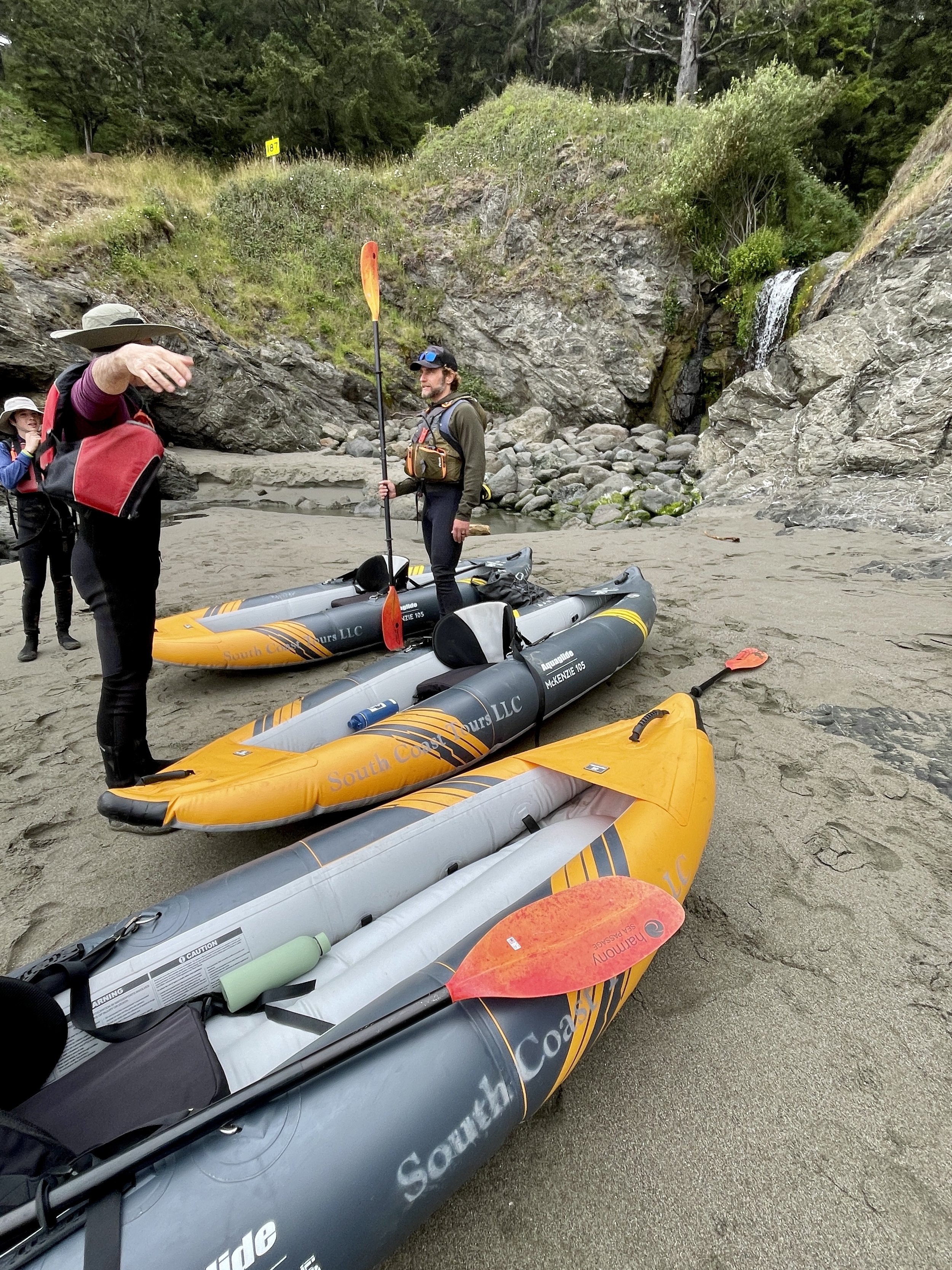 Group of people preparing to kayak on a sandy riverbank surrounded by rocks and trees.