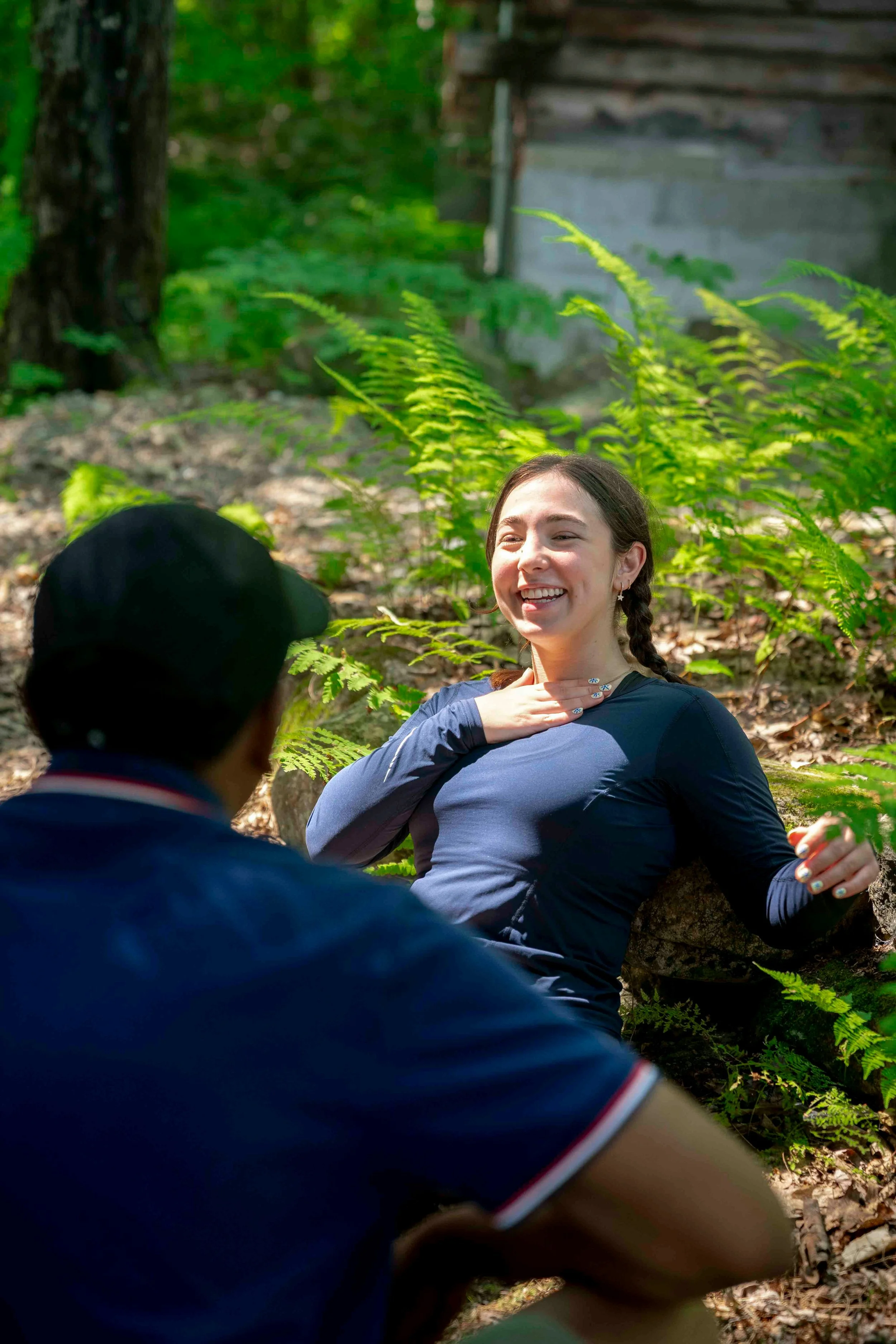 A smiling woman with braided hair sitting outdoors in a forest, with a person in a baseball cap in the foreground, and trees and a wooden structure in the background.