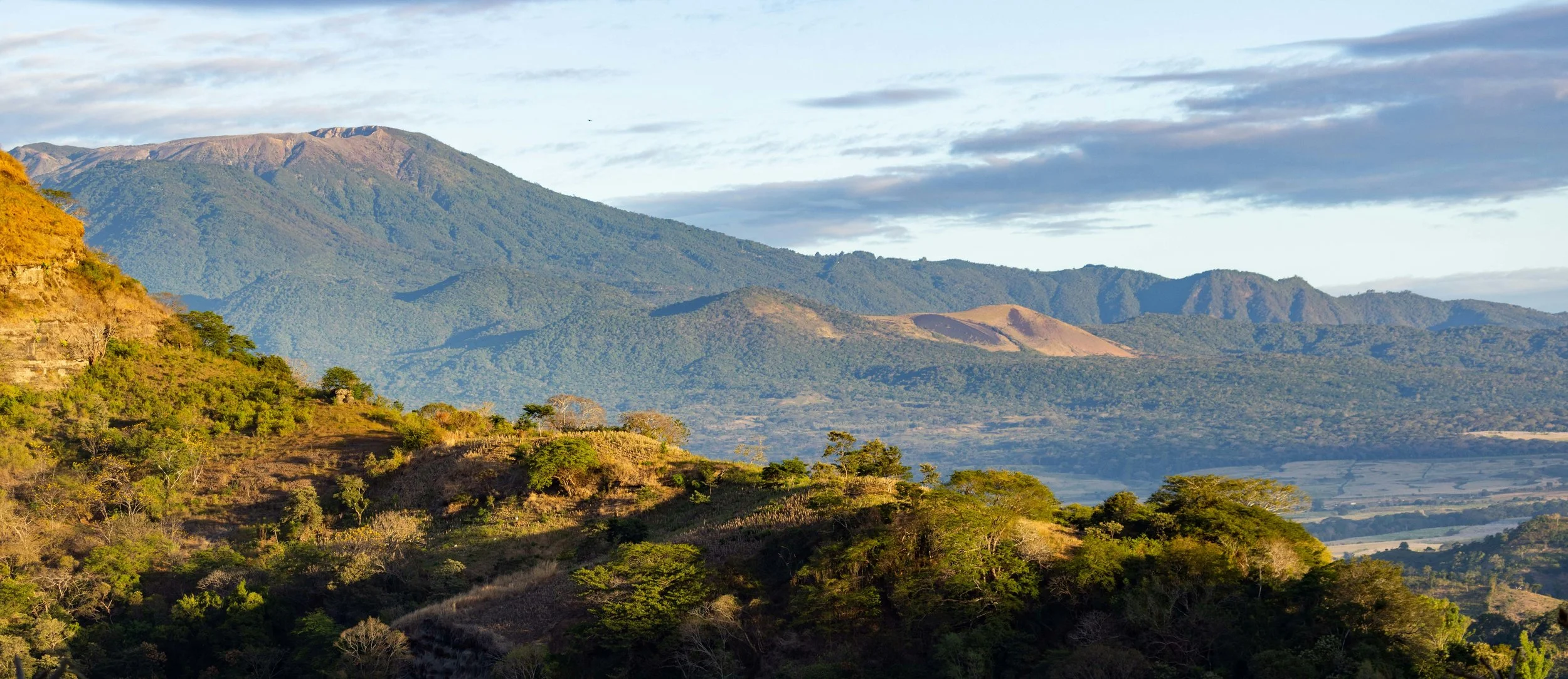Scenic landscape of mountains with green vegetation, rolling hills, and a partly cloudy sky.
