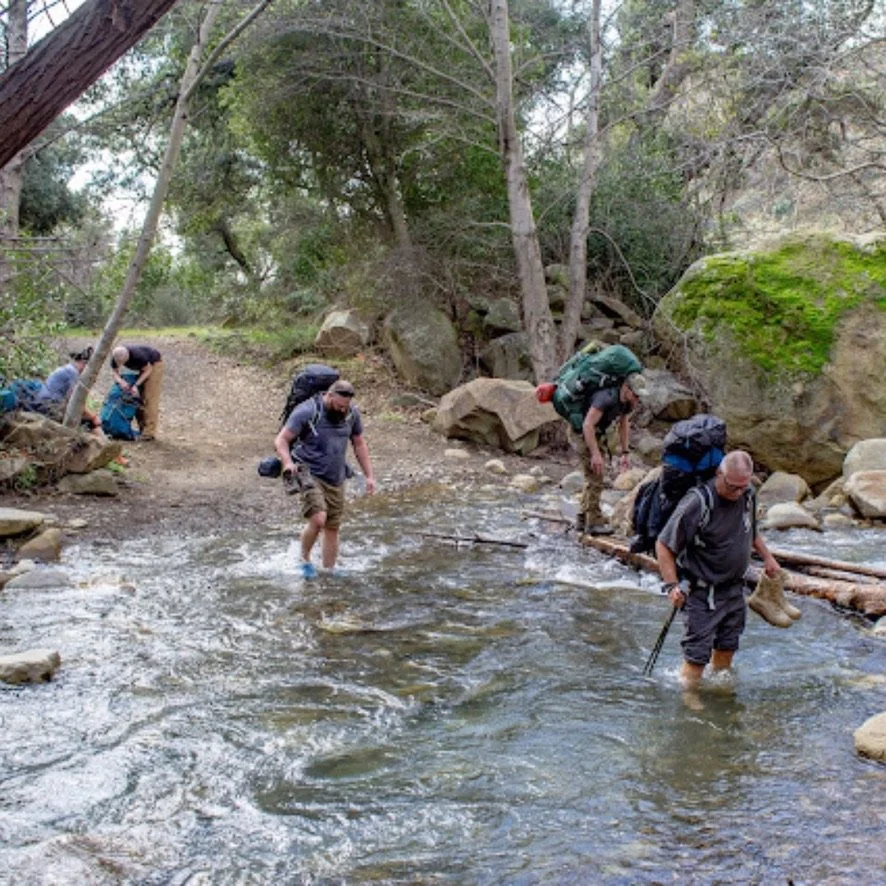 Group of hikers crossing a shallow creek on a forest trail with trees and rocks.