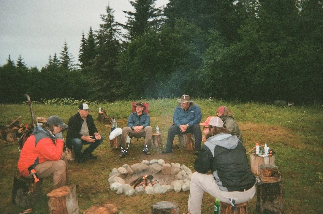 Group of people sitting in a circle around a campfire in an outdoor forest setting, with trees and grass in the background.