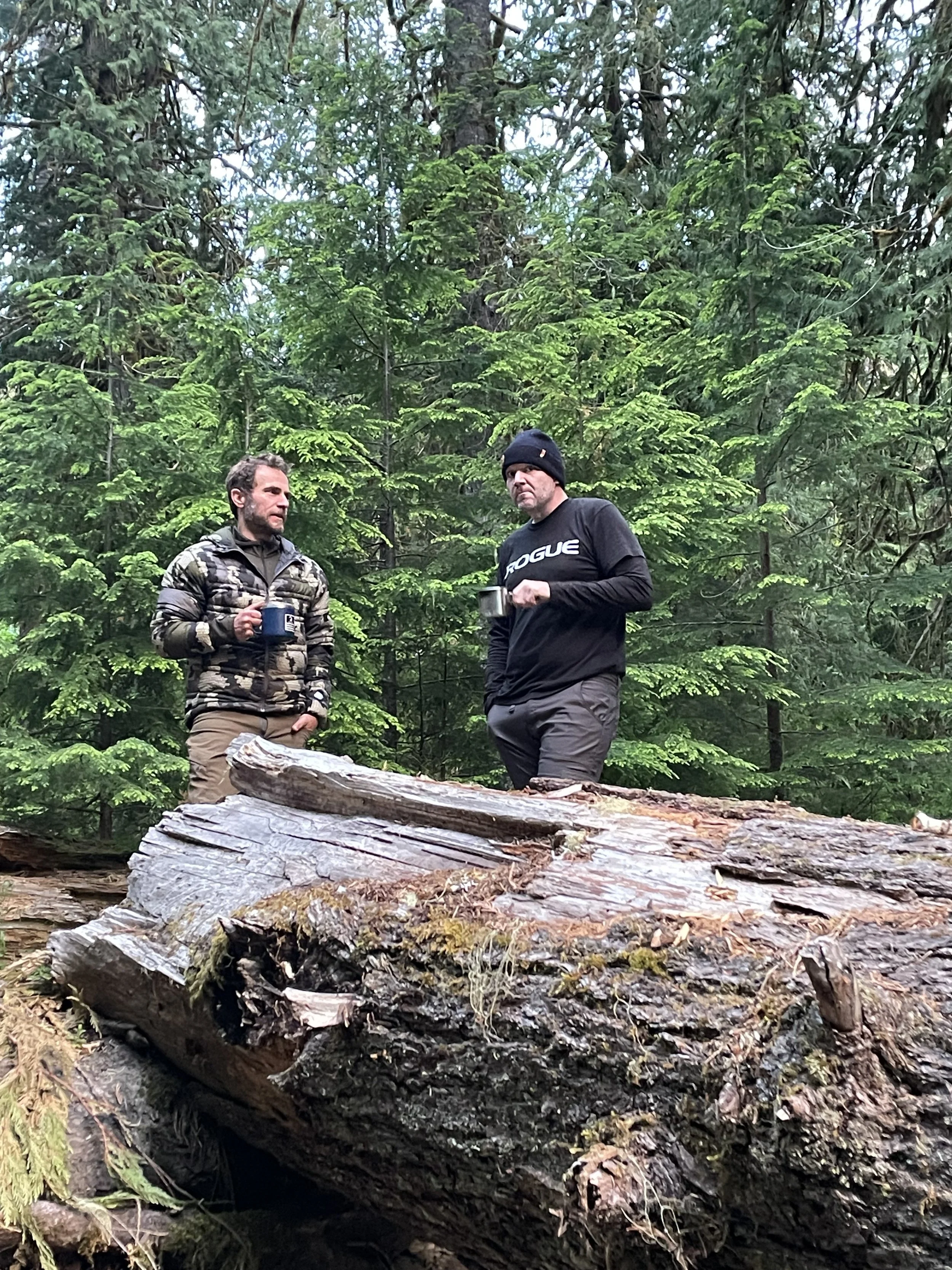 Two men standing in a forest, holding mugs of hot drinks, surrounded by tall green trees and fallen logs.