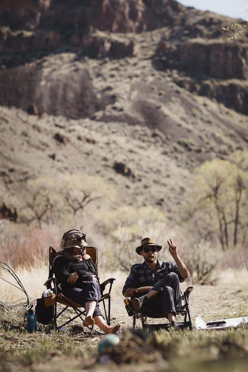 Two people sitting in camping chairs outdoors in a desert landscape with rocky hills and sparse trees, one giving a peace sign gesture.