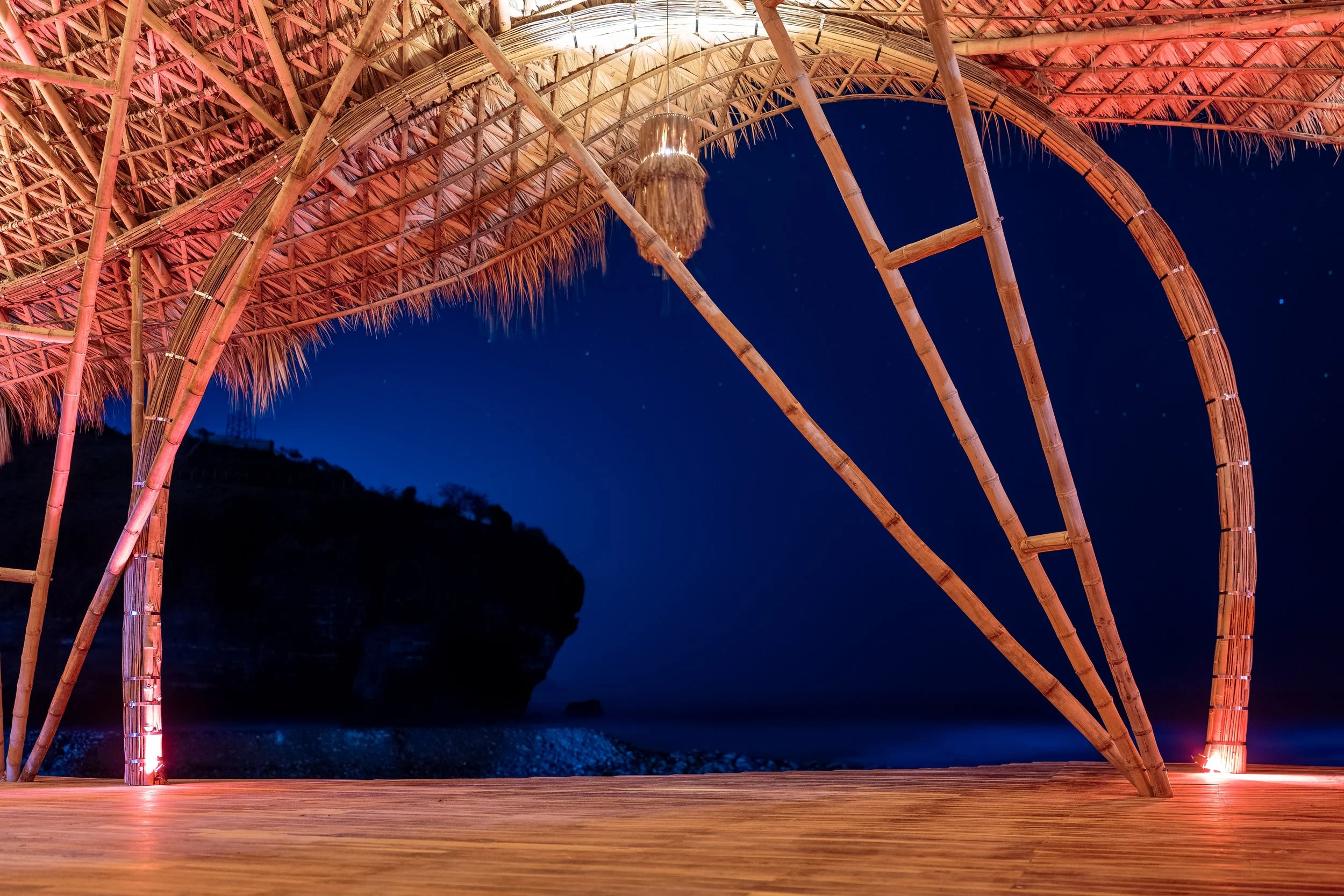 A bamboo and thatch beach hut shell at night with a view of the ocean and a rocky cliff in the distance.