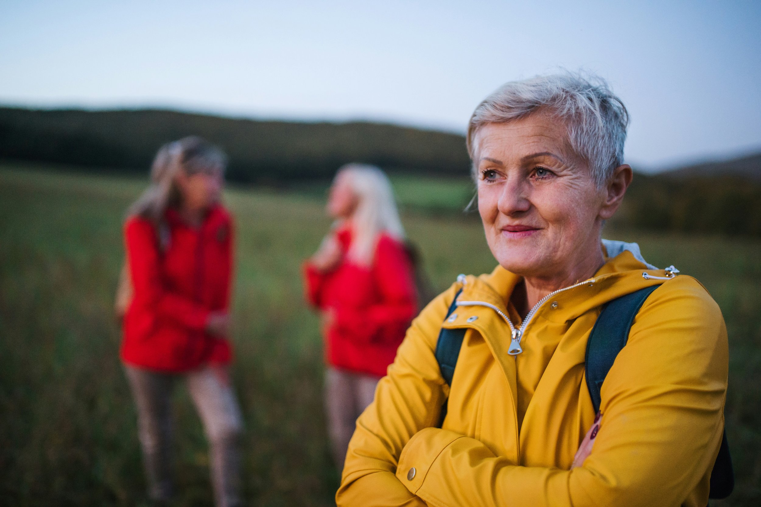 An elderly woman with short gray hair wearing a yellow jacket, standing outdoors during dusk with hills in the background. Two women in red jackets are blurred in the background.