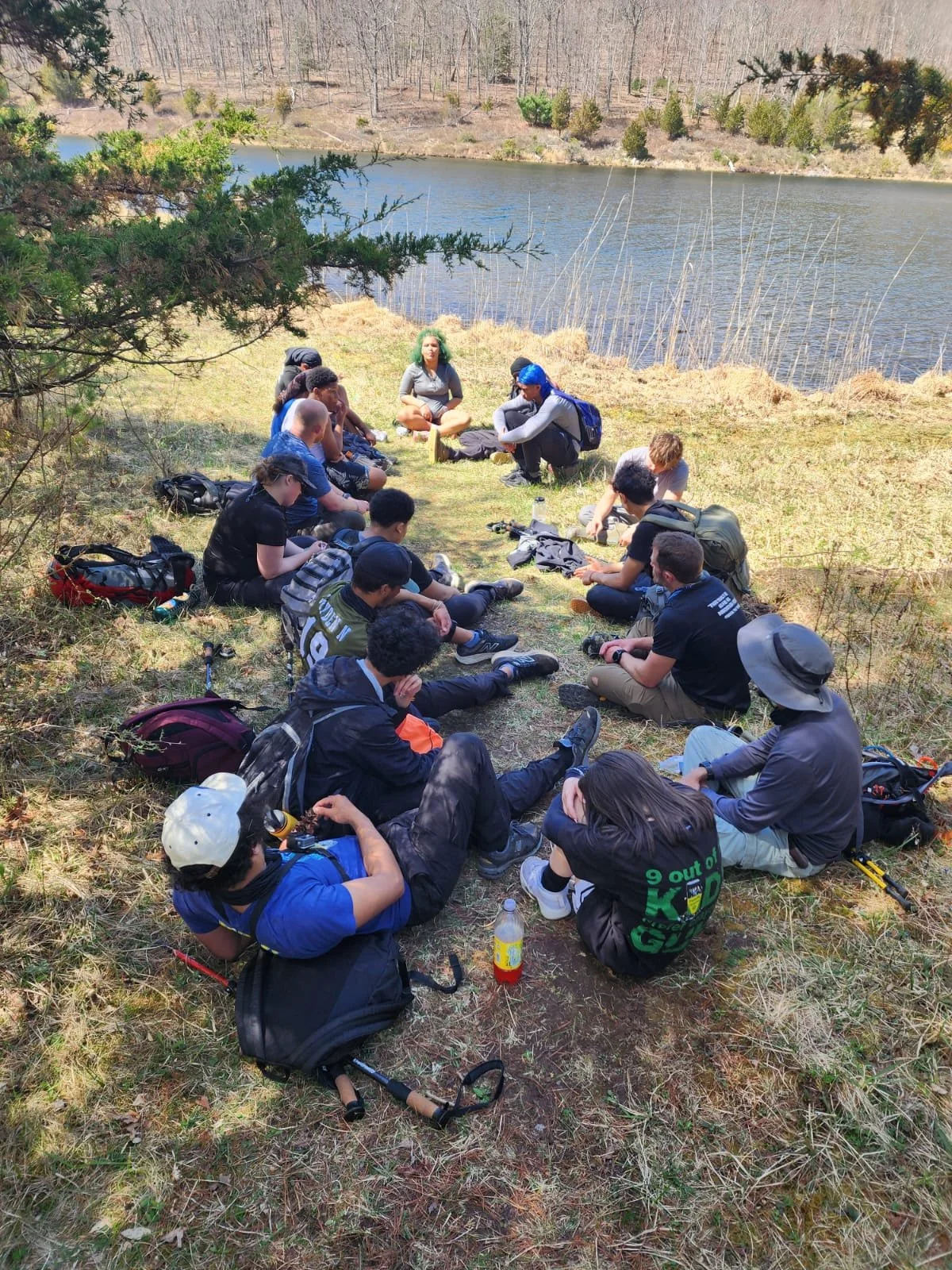 Group of people sitting in a circle outdoors near a body of water, surrounded by trees and grass.