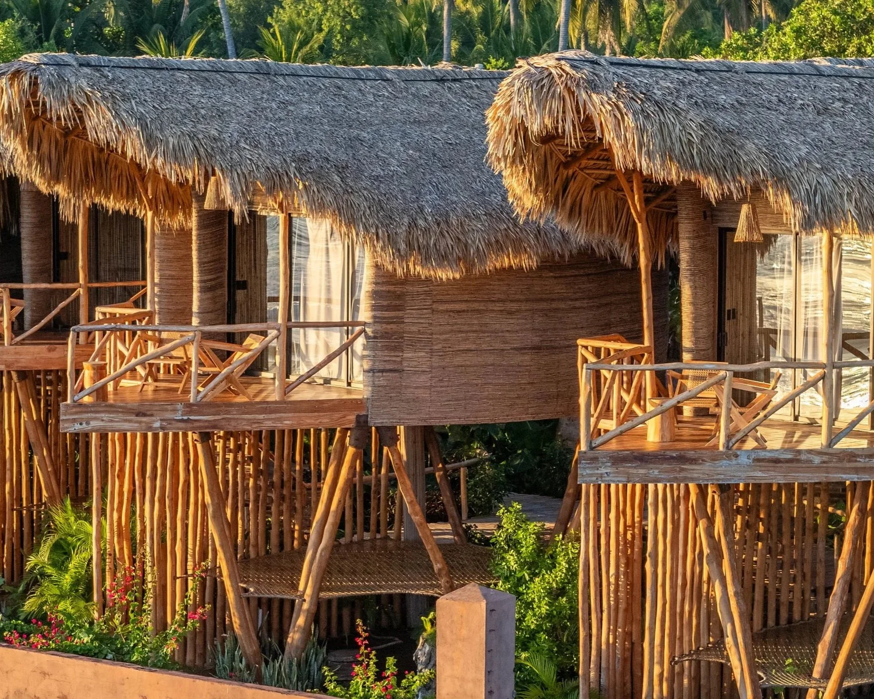 Two thatched-roof treehouse structures with wooden railings and furniture in a lush, tropical setting.