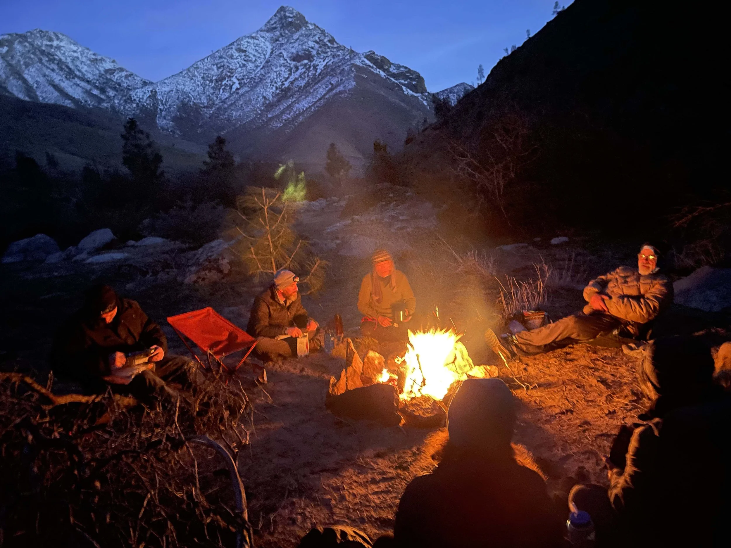 A group of people camping around a campfire in a mountainous area during dusk or nighttime, with snow-capped mountains in the background and a clear sky.