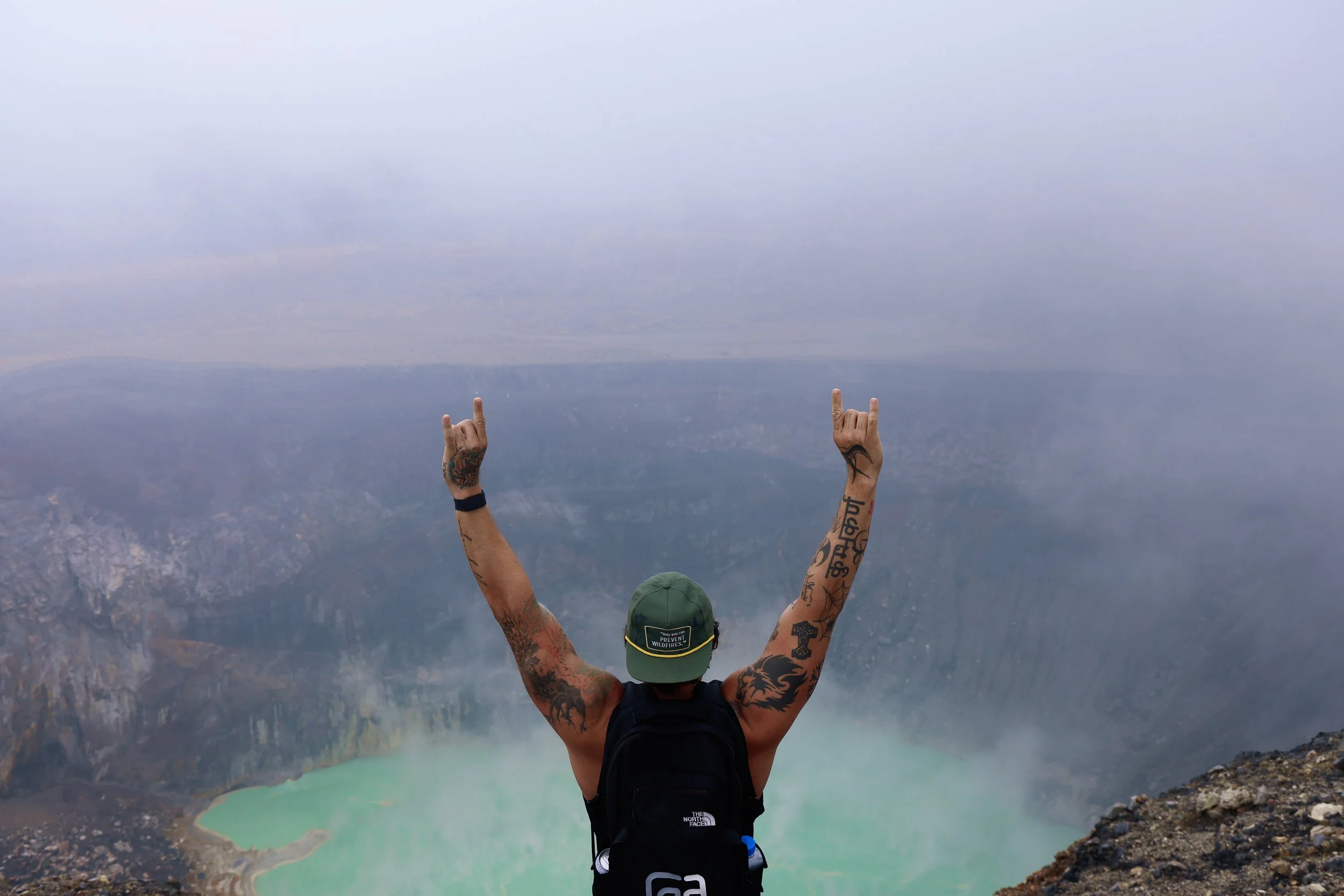 A person with tattoos and a backpack standing on a mountain, raising both arms with rock hand signs, overlooking a volcanic crater lake with cloudy sky.